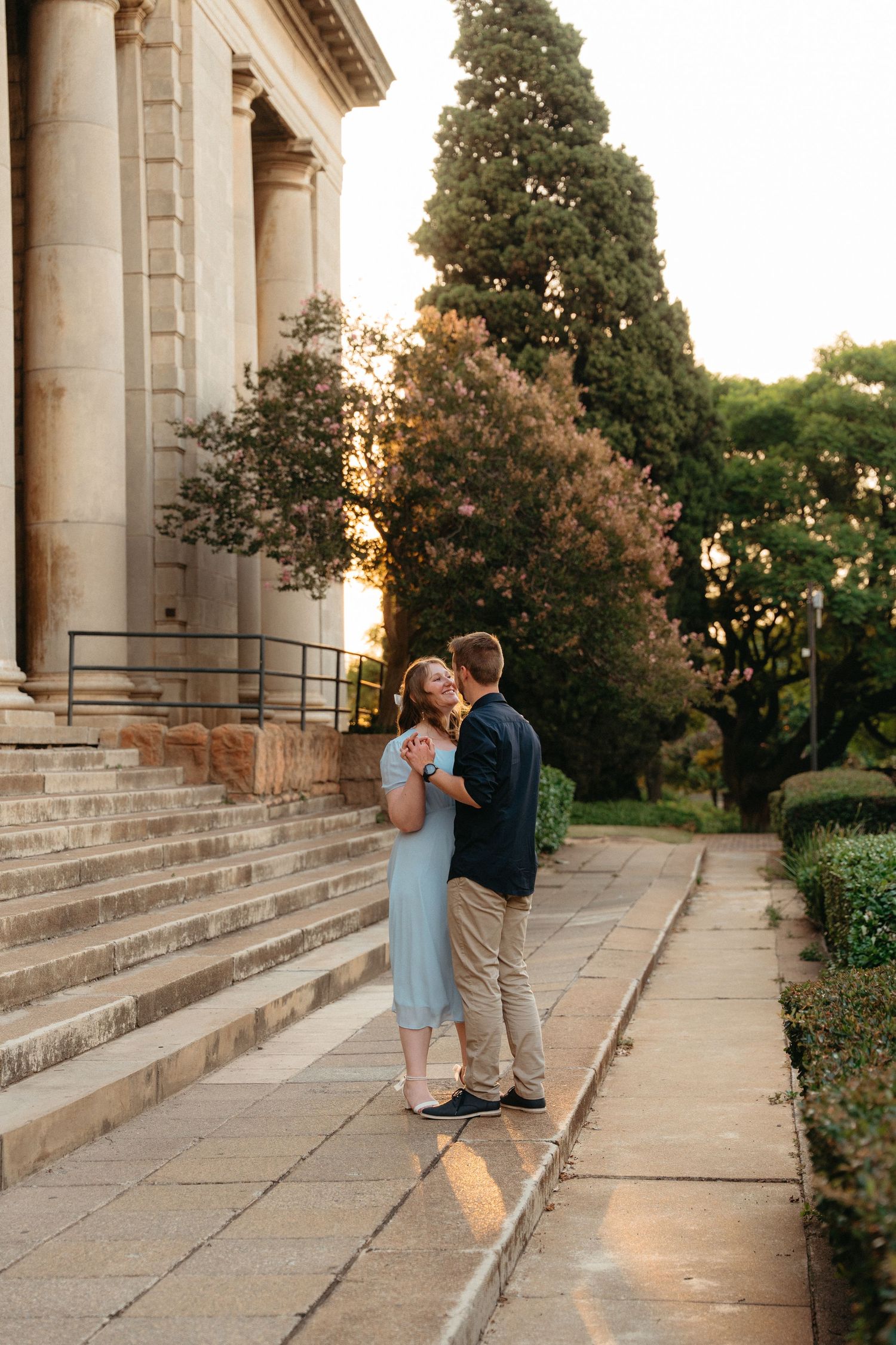 A couple embraces on stone steps outside a grand building with columns during golden hour sunset.