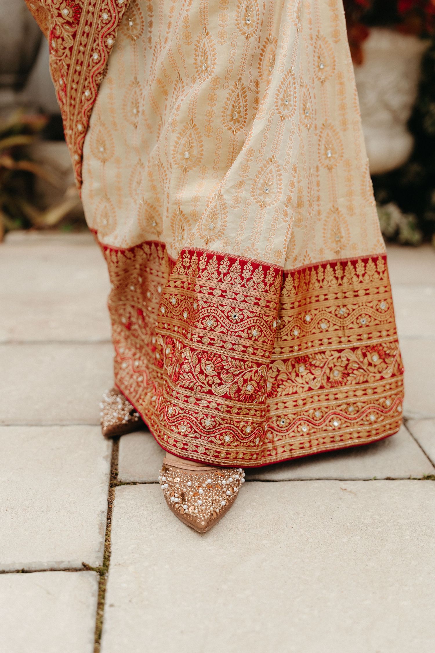 Close-up details of an ornate red and gold embroidered Indian wedding lehenga skirt hem on stone pavement.