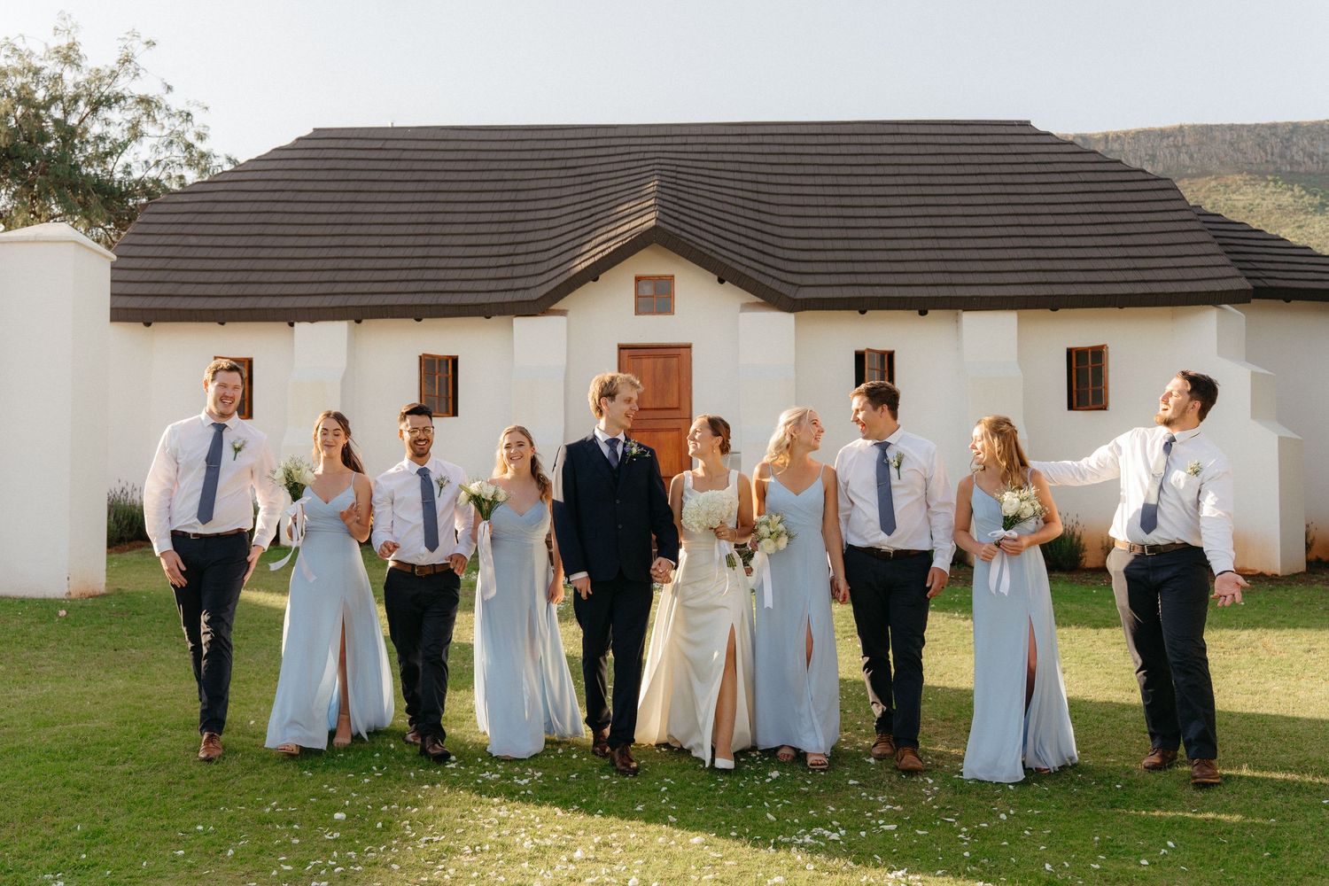 Wedding party in light blue dresses and dark suits poses in line formation outside white building.