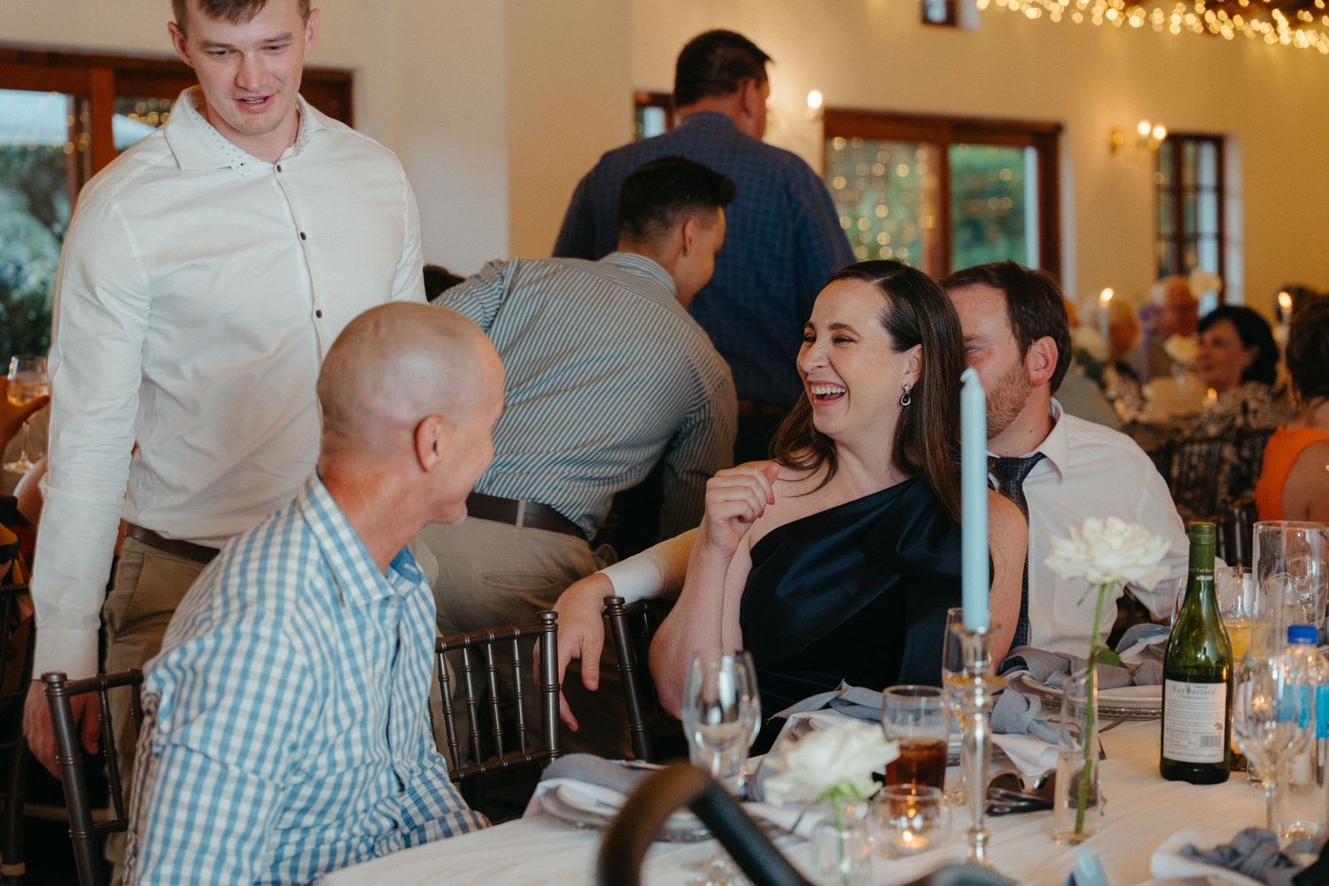 A group of guests seated at long dining tables in a festive indoor venue with string lights overhead.