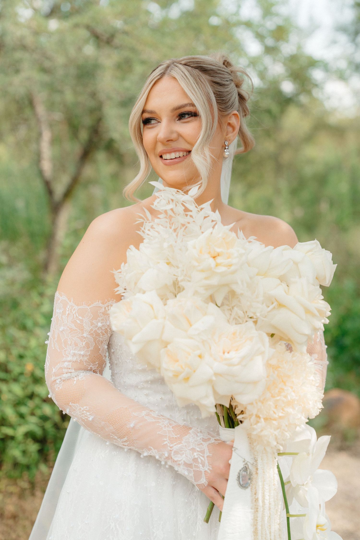 Smiling bride in lace off-shoulder gown holding large white peony bouquet in a bush setting in Pretoria