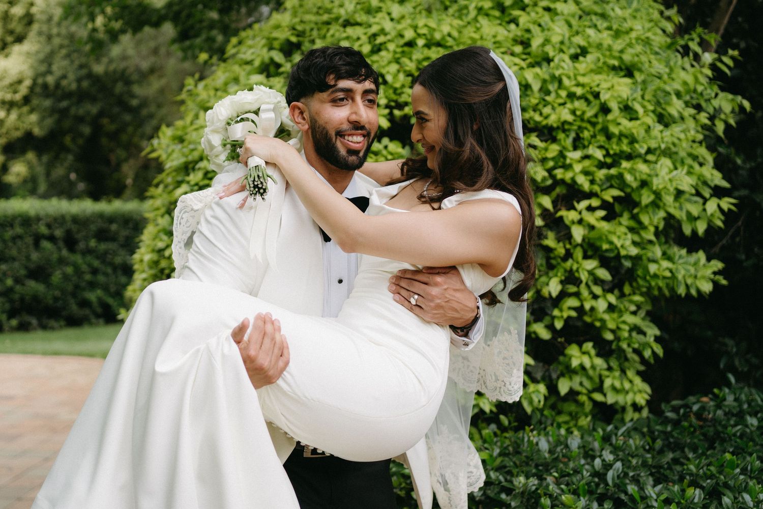 A joyful couple shares an intimate moment in their wedding attire against lush green foliage in a garden setting.