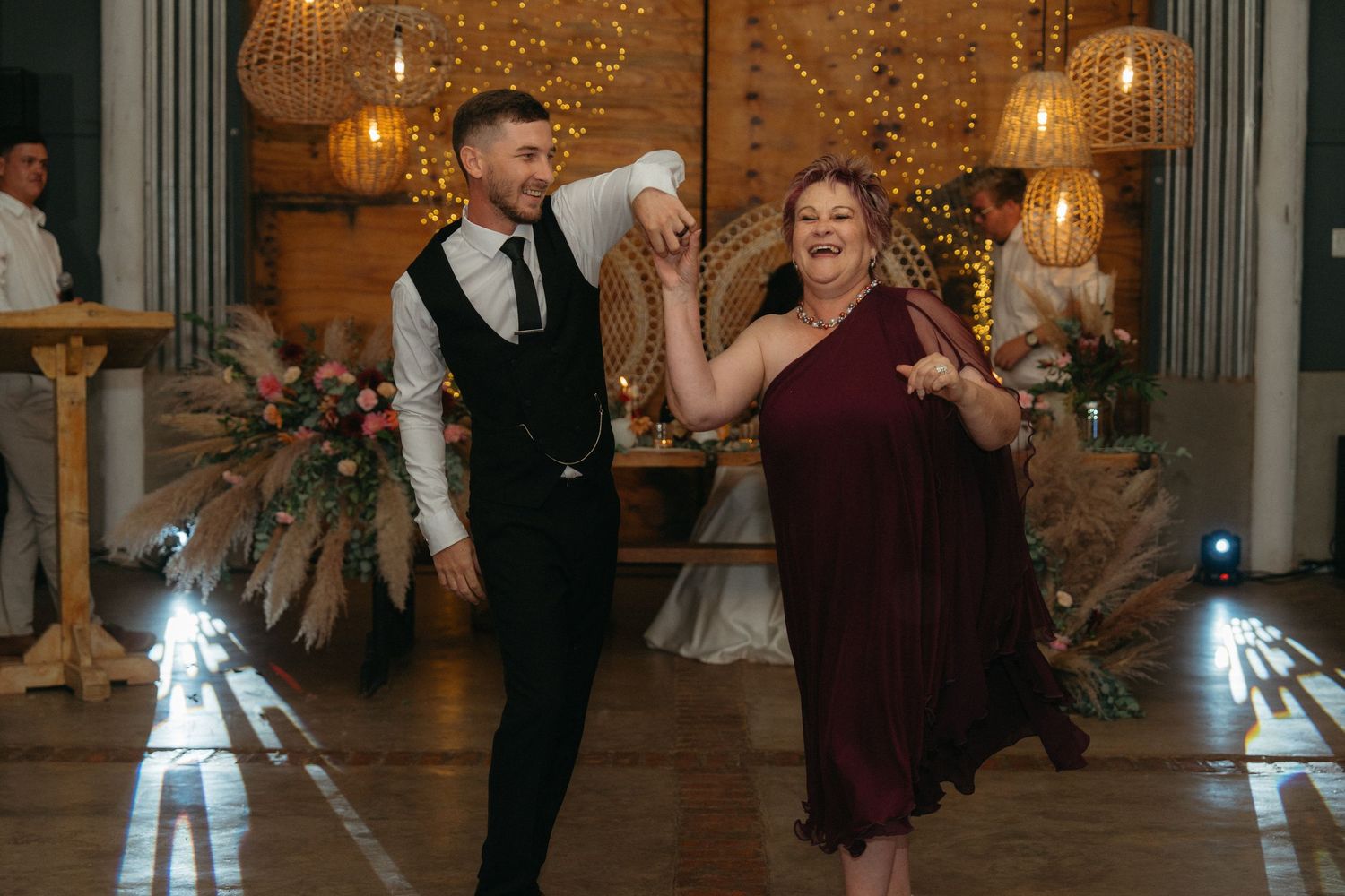 Wedding guests dancing and celebrating at an indoor reception with decorative lighting and hanging lamps.