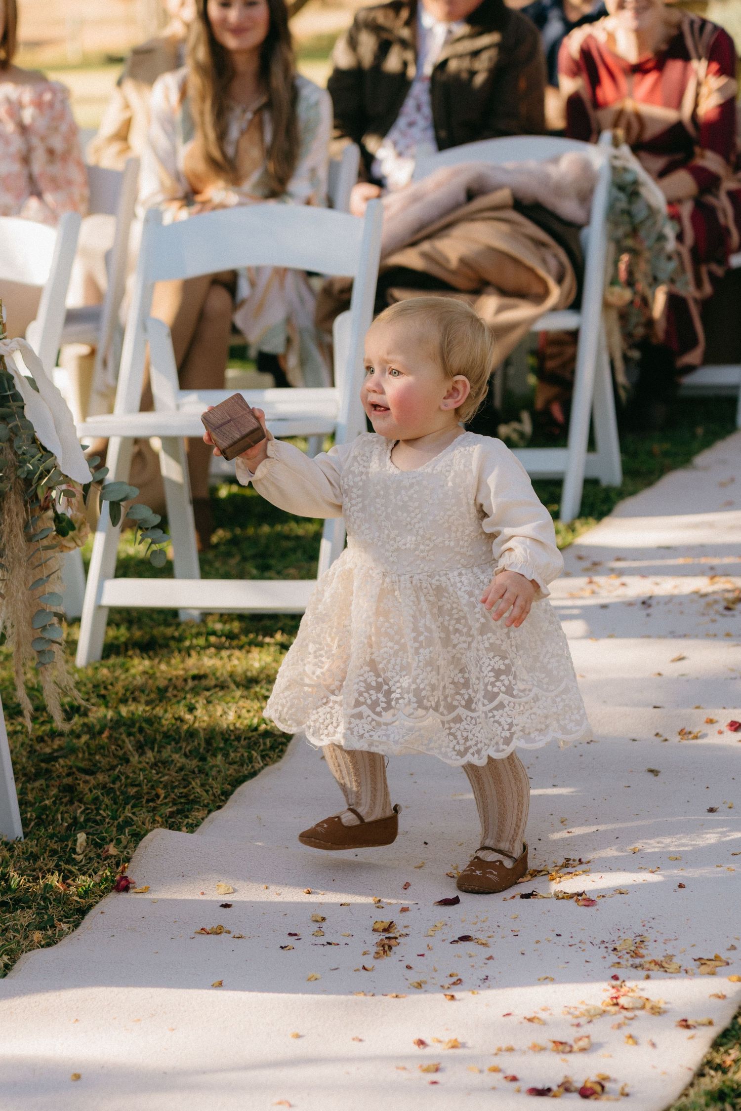 Small child in white lace dress walking down aisle scattered with flower petals.