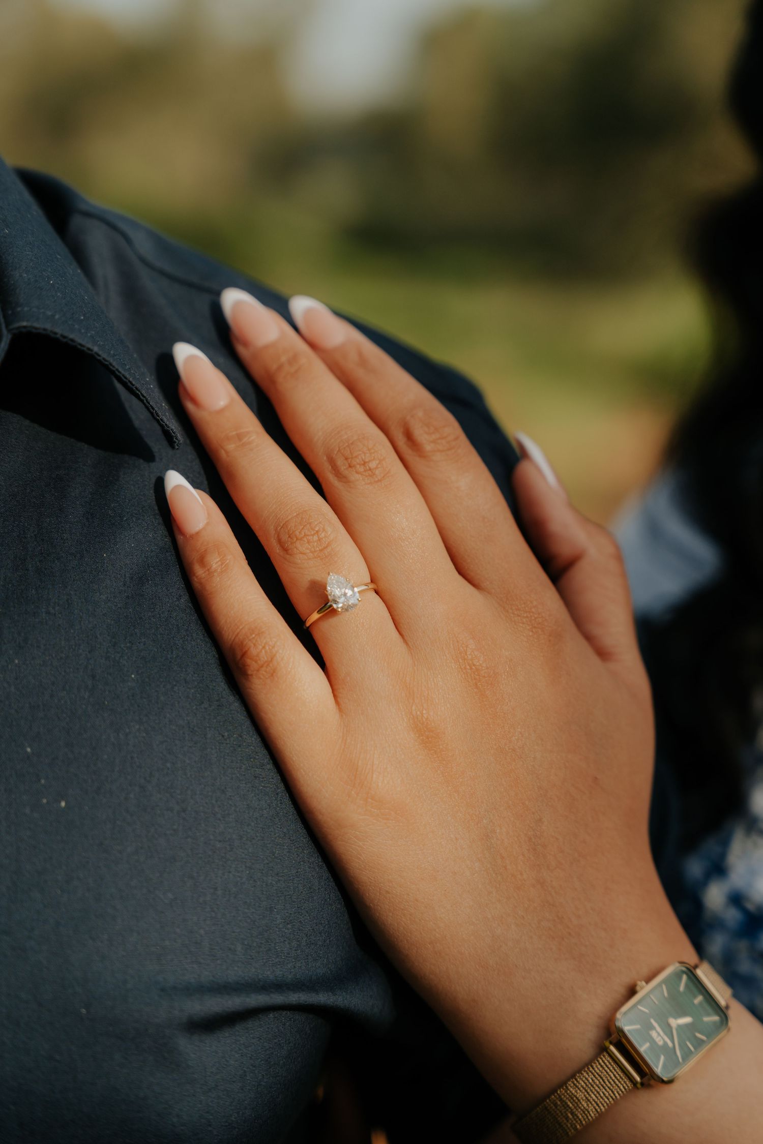 A hand wearing an engagement ring rests tenderly against a black suit jacket in an intimate engagement photo.