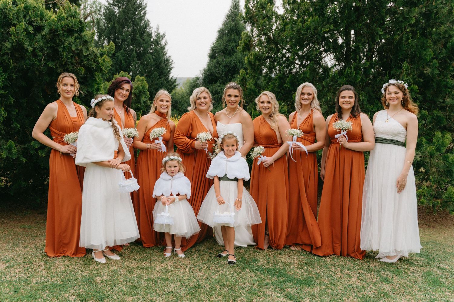 Wedding party in alternating white and rust-colored dresses pose together on grass.