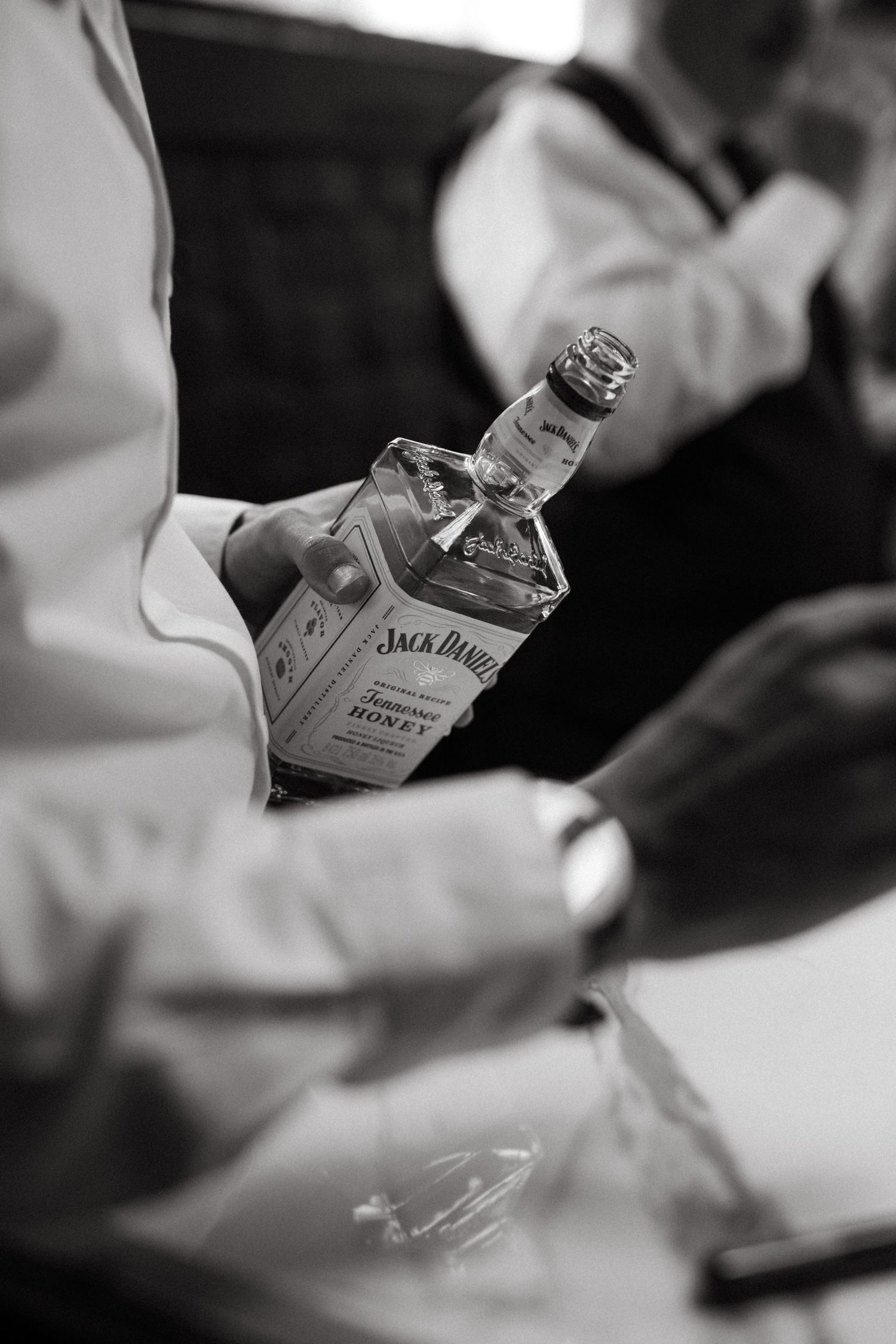 A black and white close-up shot of a Jack Daniel's whiskey bottle being held by hands in white sleeves.