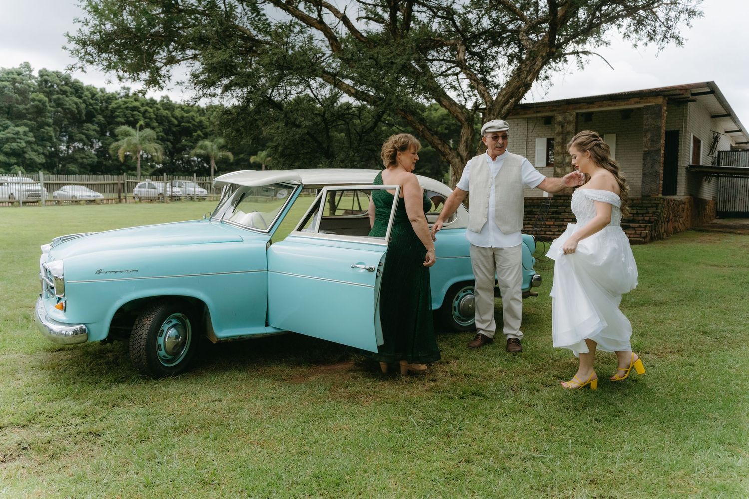 Wedding party posing beside vintage light blue car in outdoor countryside setting.