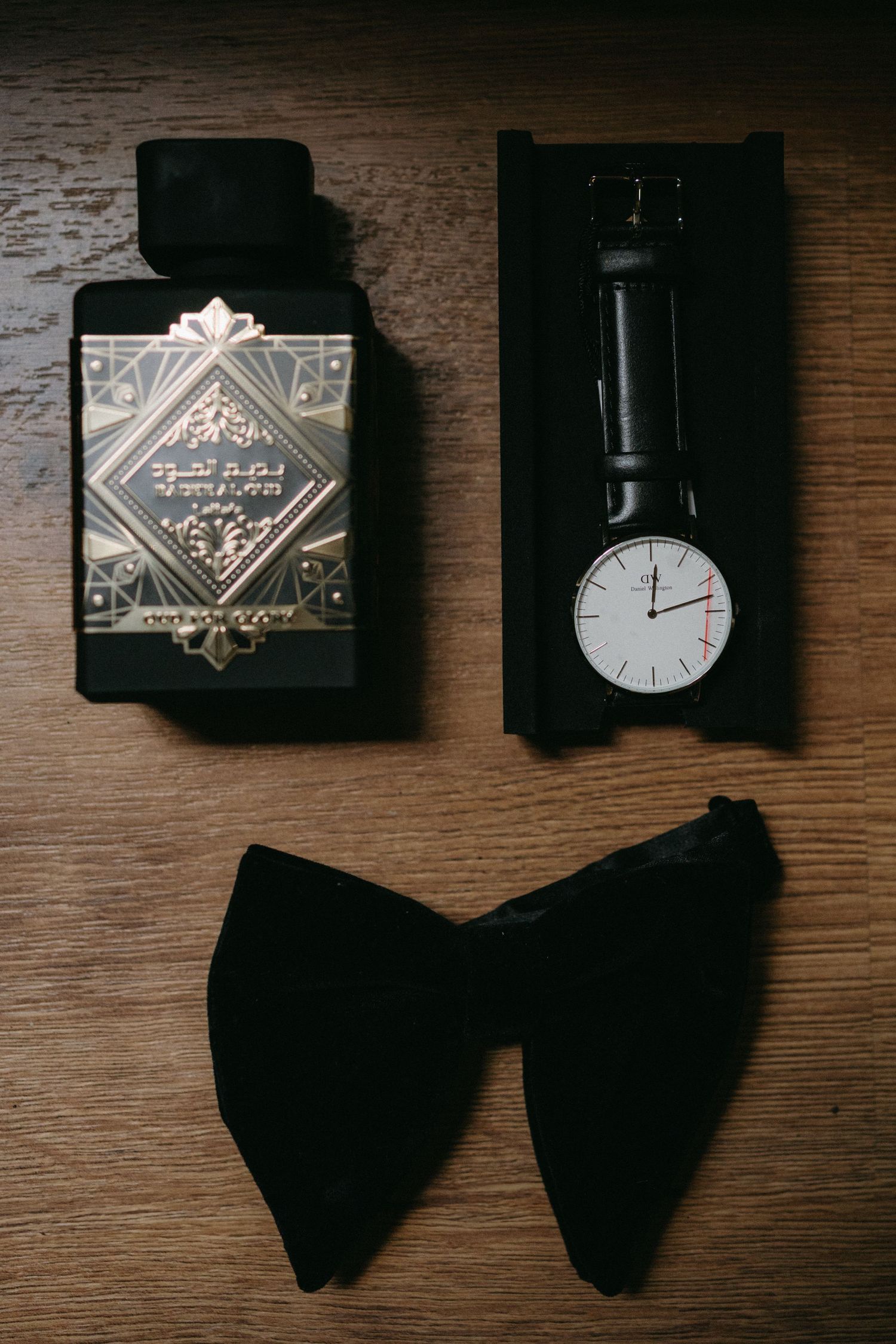 Wedding accessories including black bow tie, perfume bottle and wristwatch arranged on wooden surface.