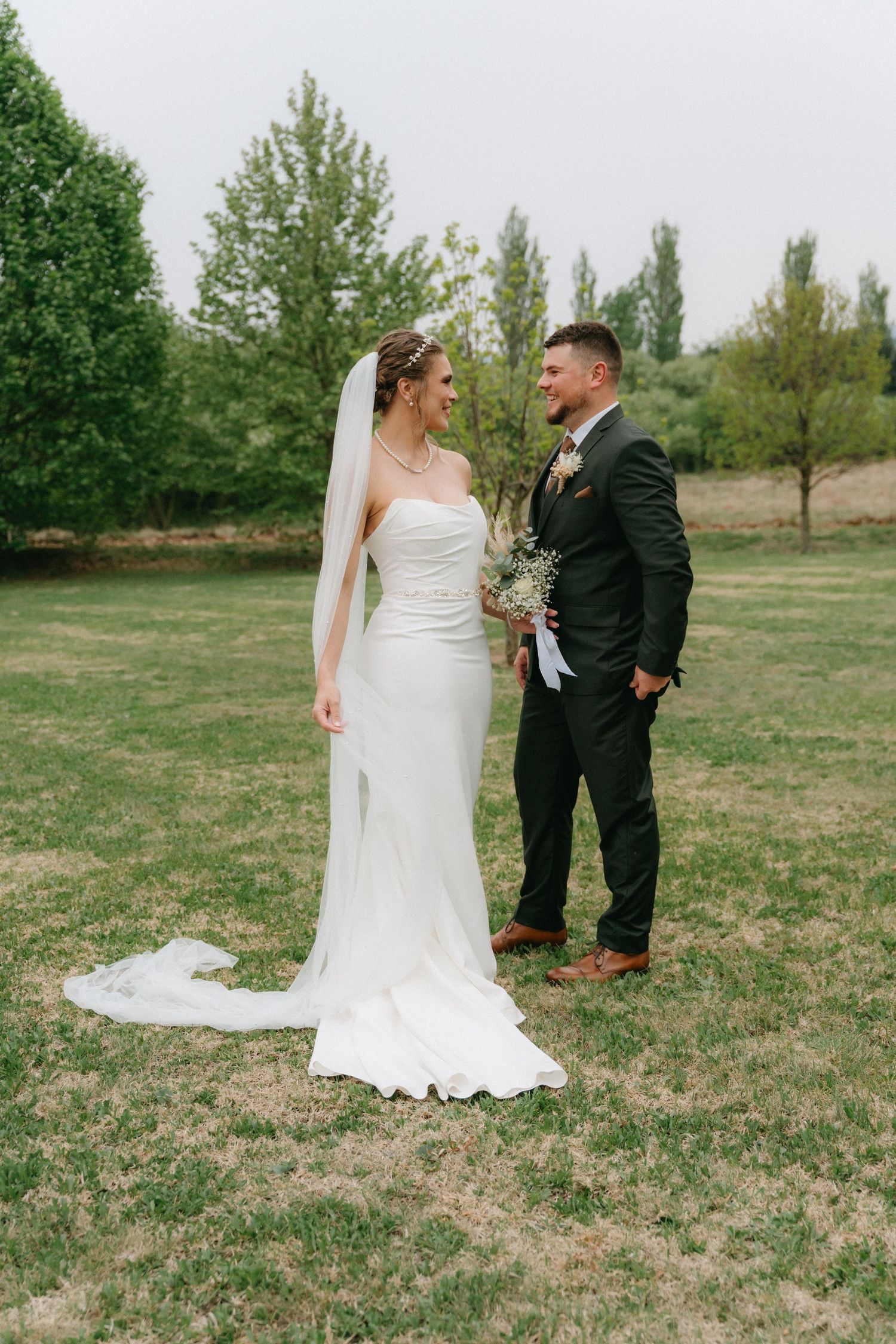 Wedding couple poses together on a grassy field with trees in the background.