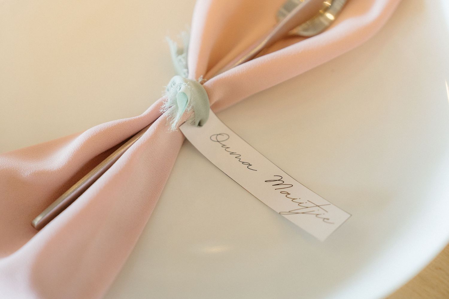 A peach-colored fabric napkin with an elegant name tag tied around it sits on a white plate.