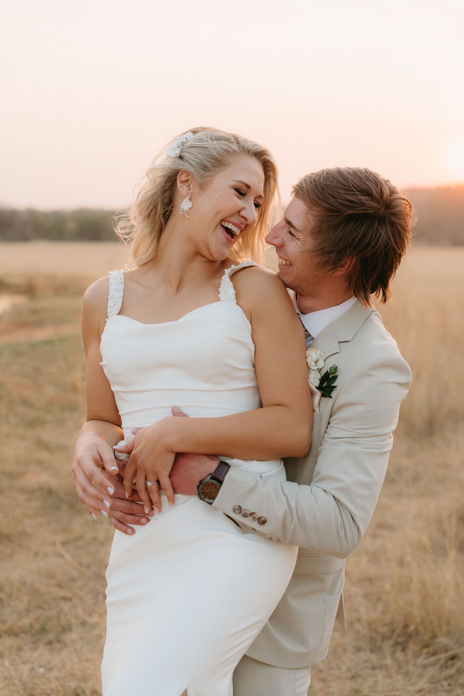Happy couple embracing and laughing together in a sunlit field during their wedding photoshoot.