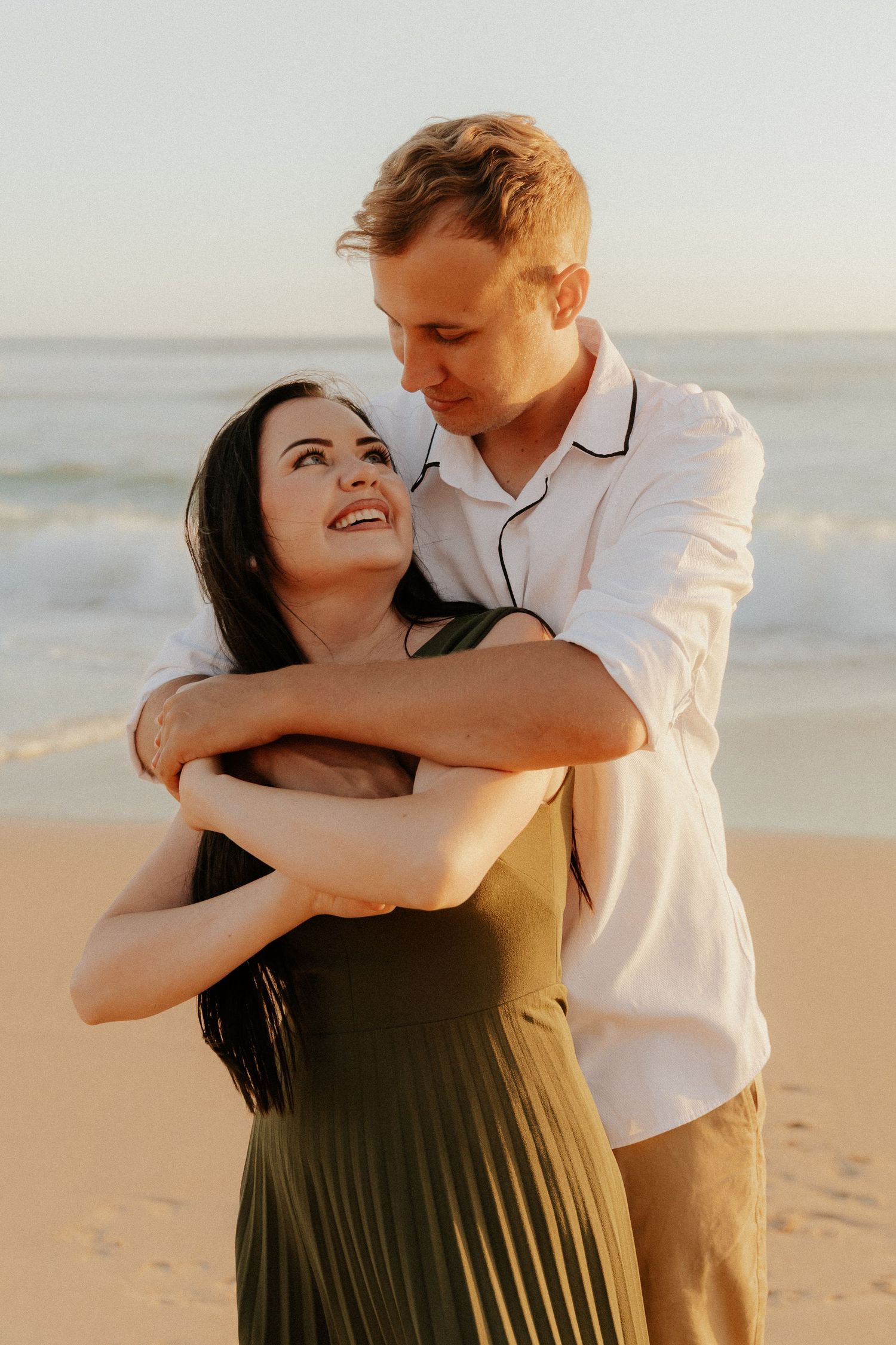 A couple embraces and laughs together on a sunny beach at sunset, creating a joyful and romantic moment.