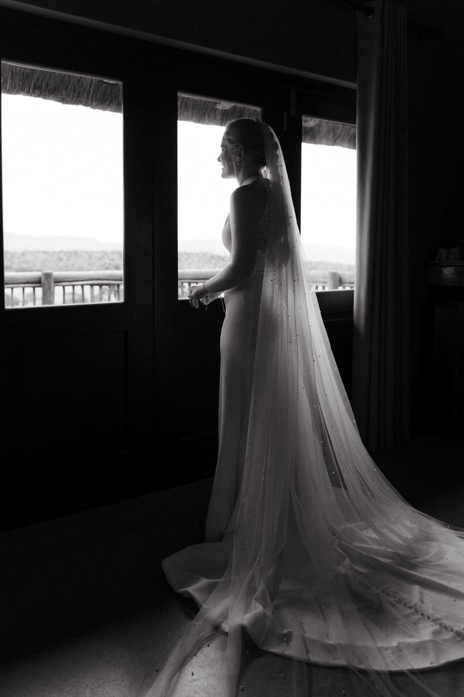 Silhouette of wedding dress by window creates dramatic black and white composition.