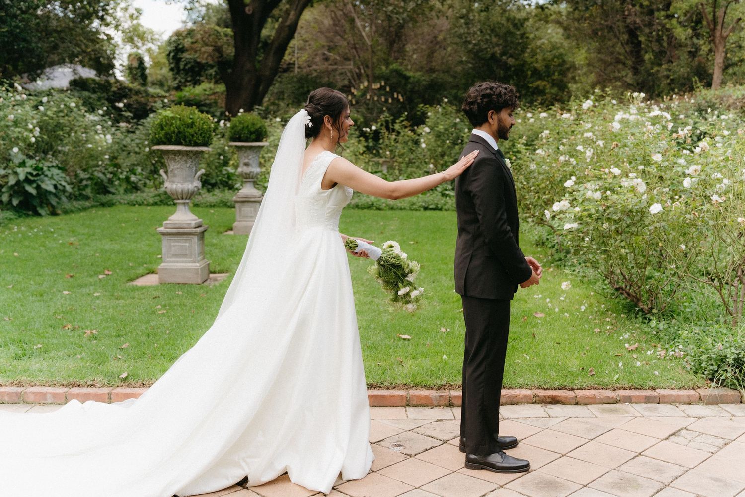 A couple shares their first look moment in a garden setting with white daisies and a stone pedestal.