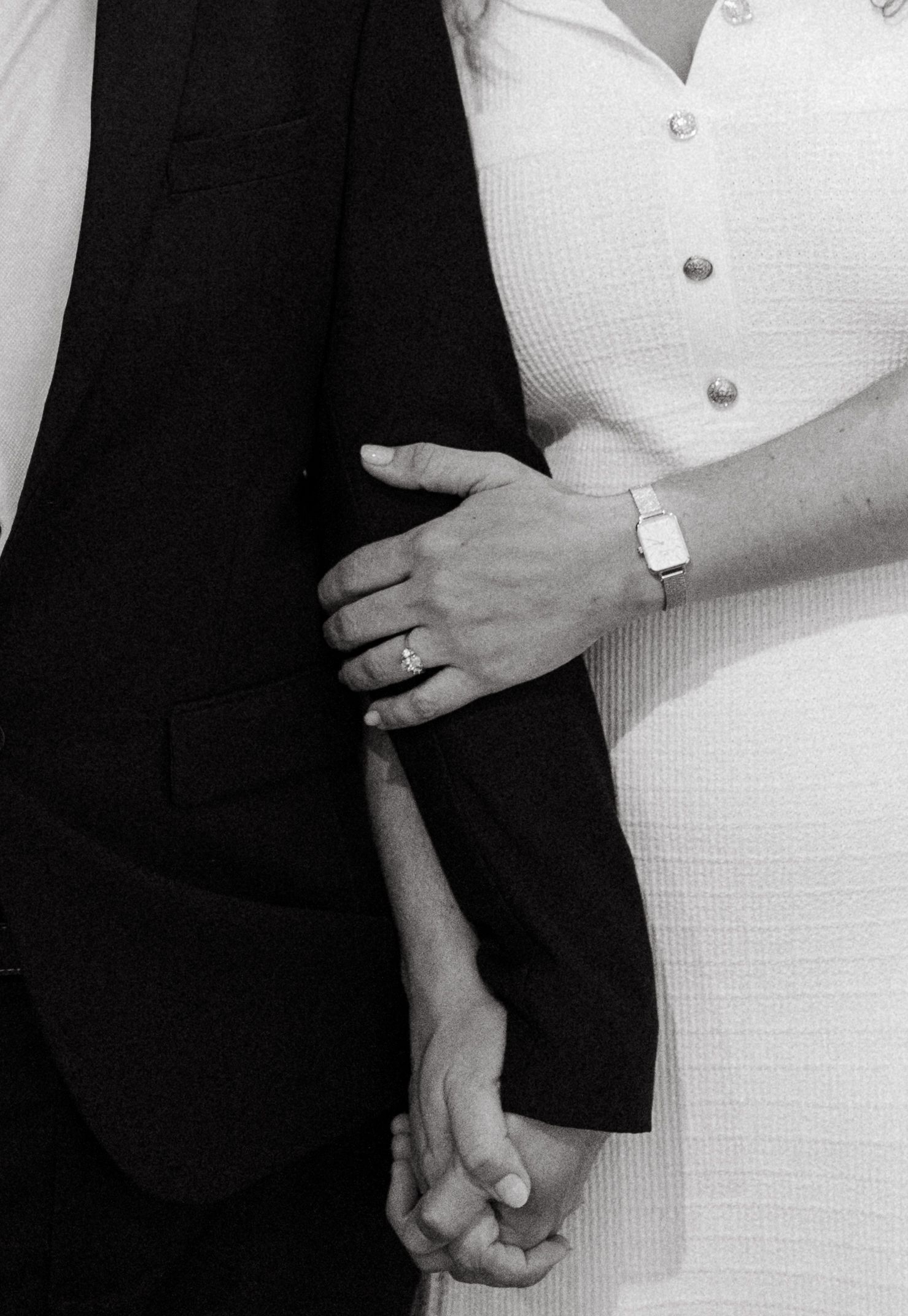Close-up black and white photo of couple holding hands showing wedding rings and formal attire.