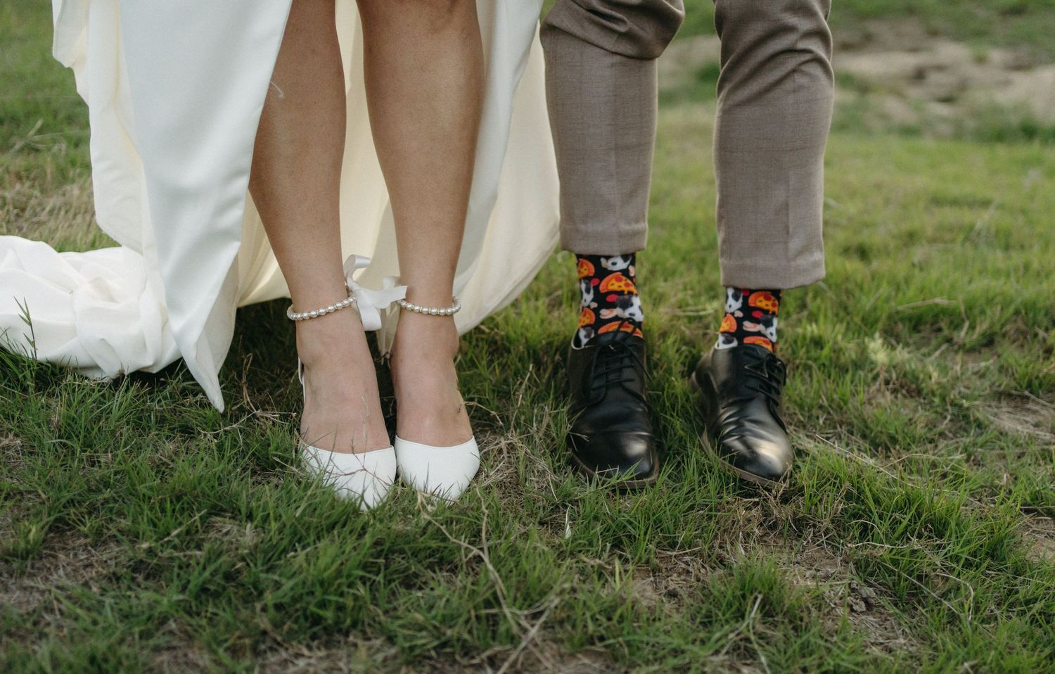 Wedding couple shows off their contrasting footwear with colorful socks and elegant shoes on grassy ground.