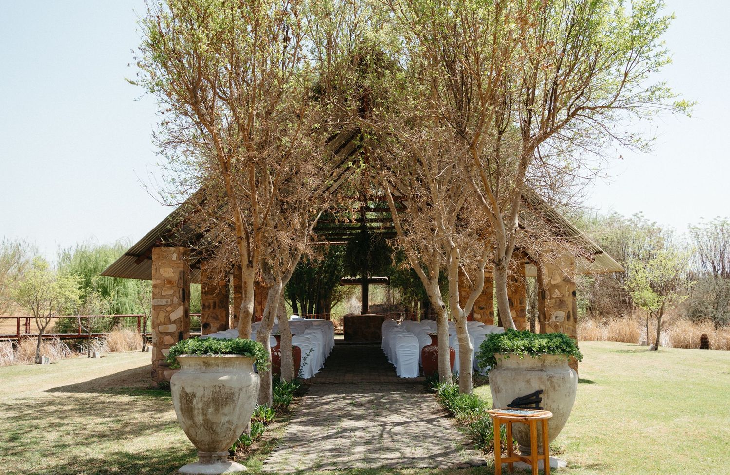 Rustic outdoor wedding venue with wooden chapel structure and decorative potted plants along the entrance path.