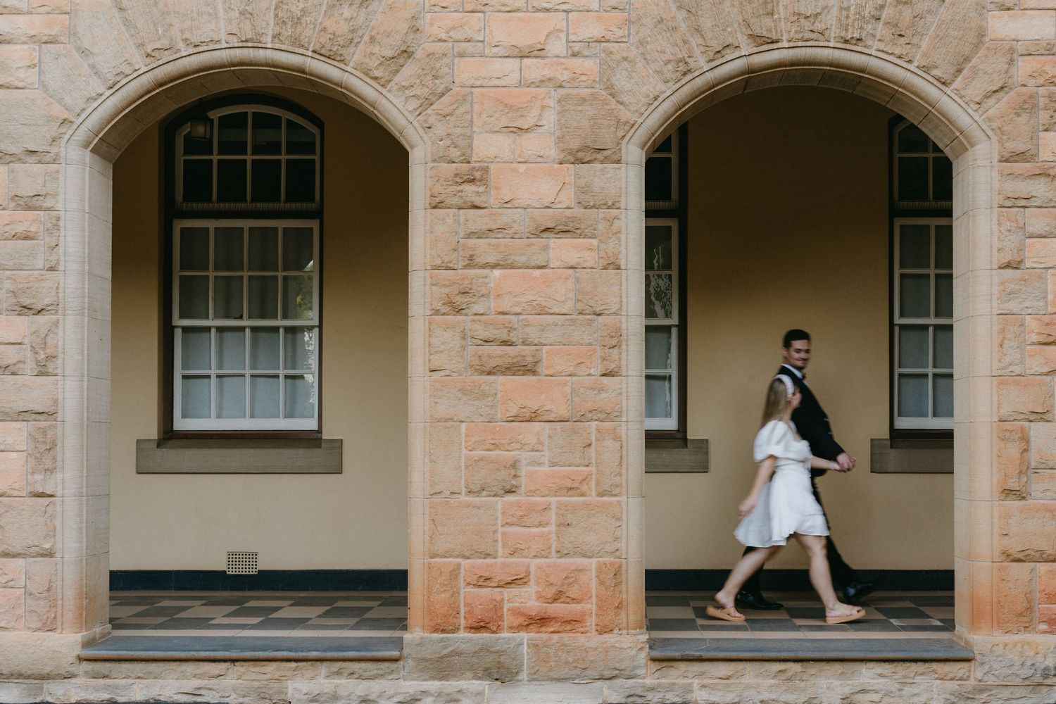 A couple walks through stone archways of a historic building with sandstone walls and arched windows.