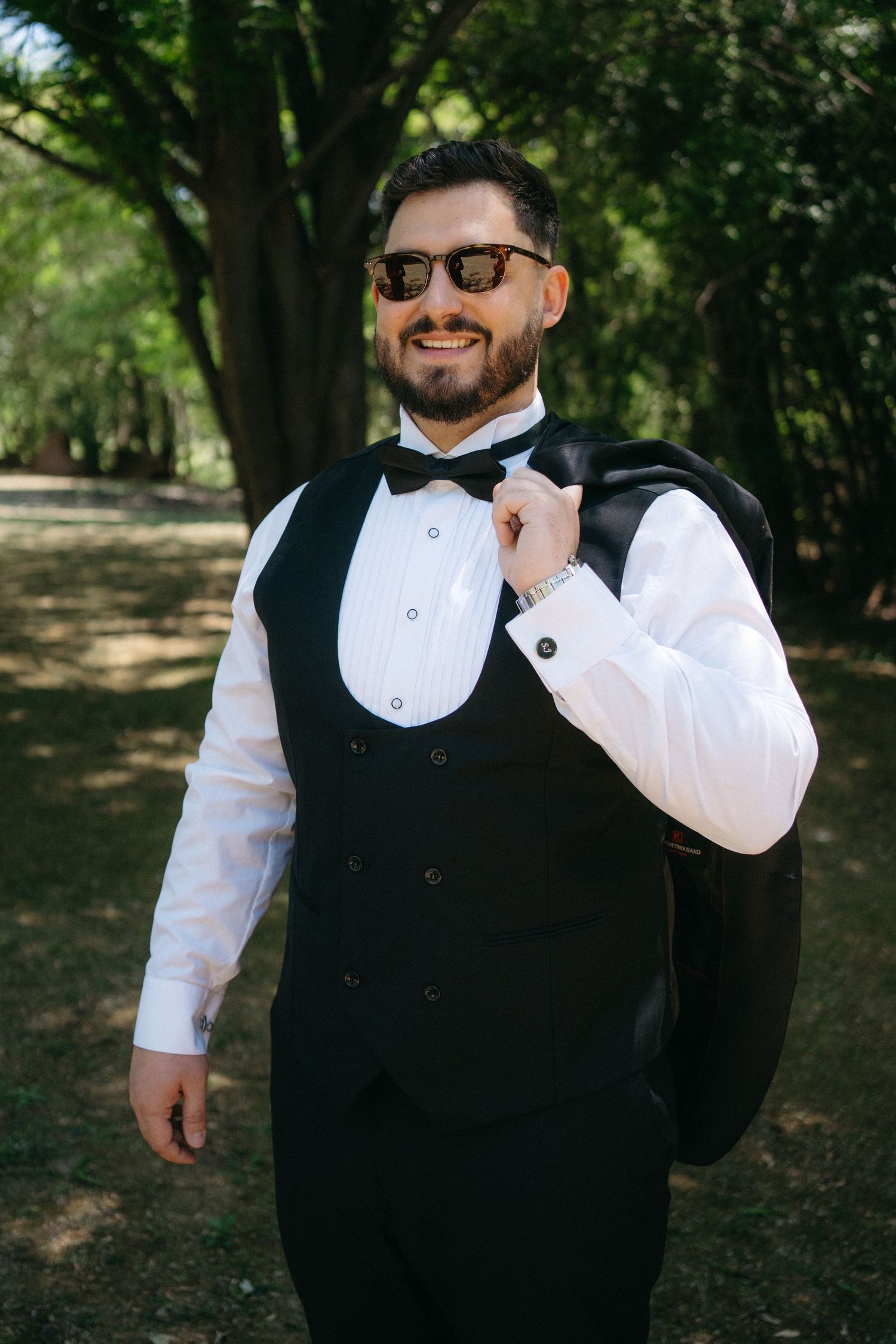 A person in formal attire with a black vest and bowtie smiles while standing outdoors among trees on a sunny day.
