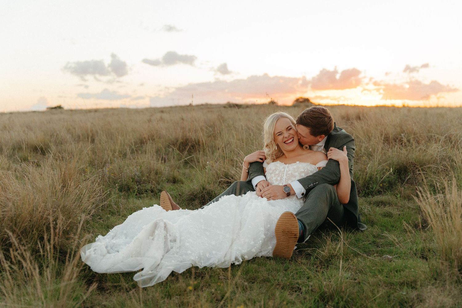 Bride and groom sitting in golden grass field at sunset, laughing and embracing in wedding attire.