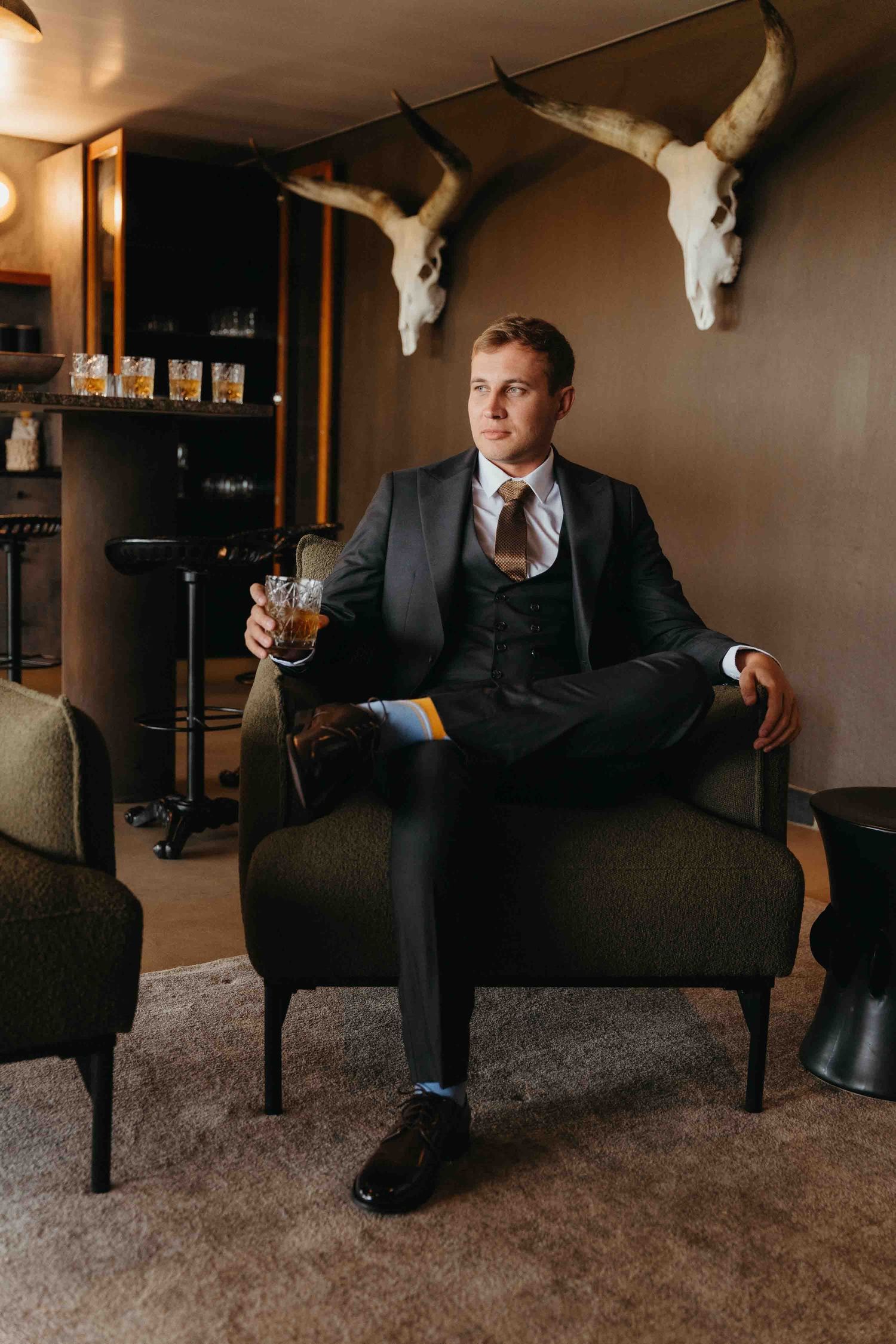 A well-dressed man in a dark suit sits in an armchair holding whiskey, with animal skull decor on the wall behind him.