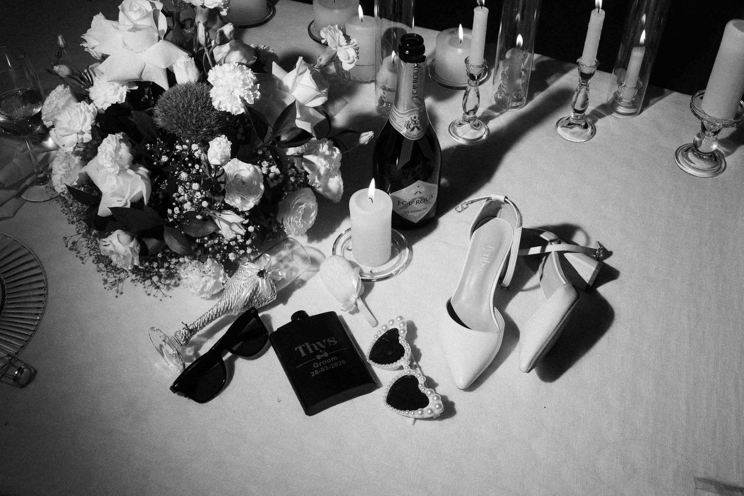 Black and white wedding flat lay with bridal heels, bouquet, champagne, candles, and perfume on a table.