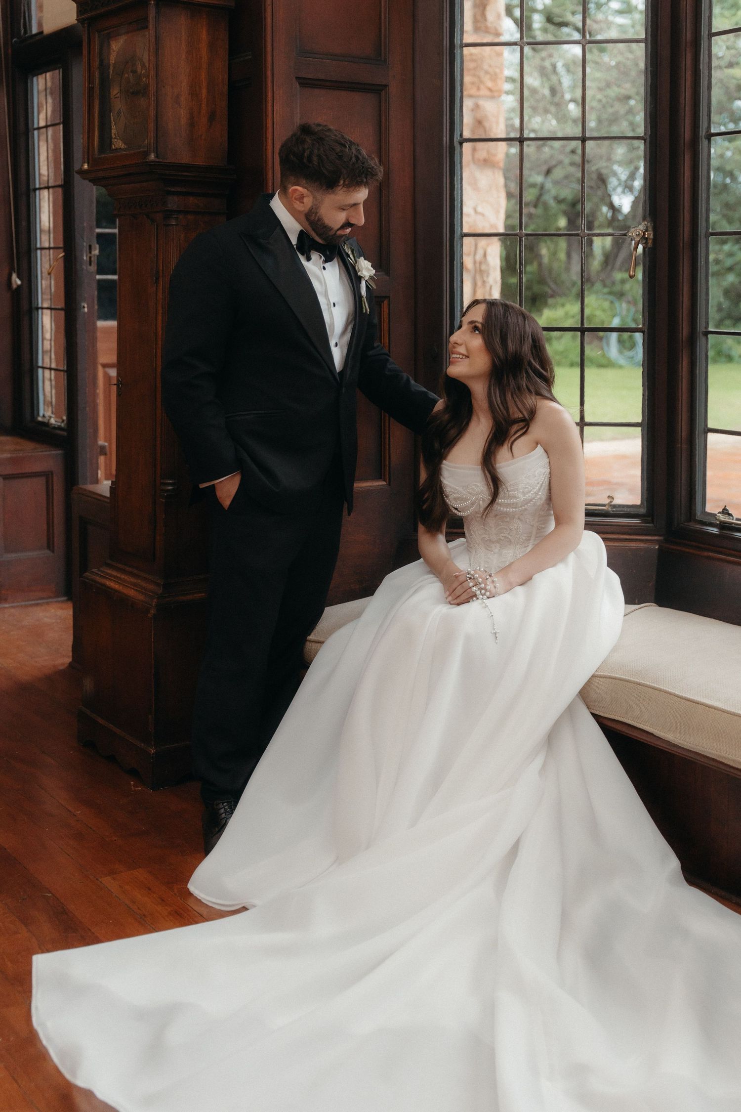 Bride in white ballgown sits gazing up at groom in black tuxedo beside tall windows in elegant wood-paneled room.