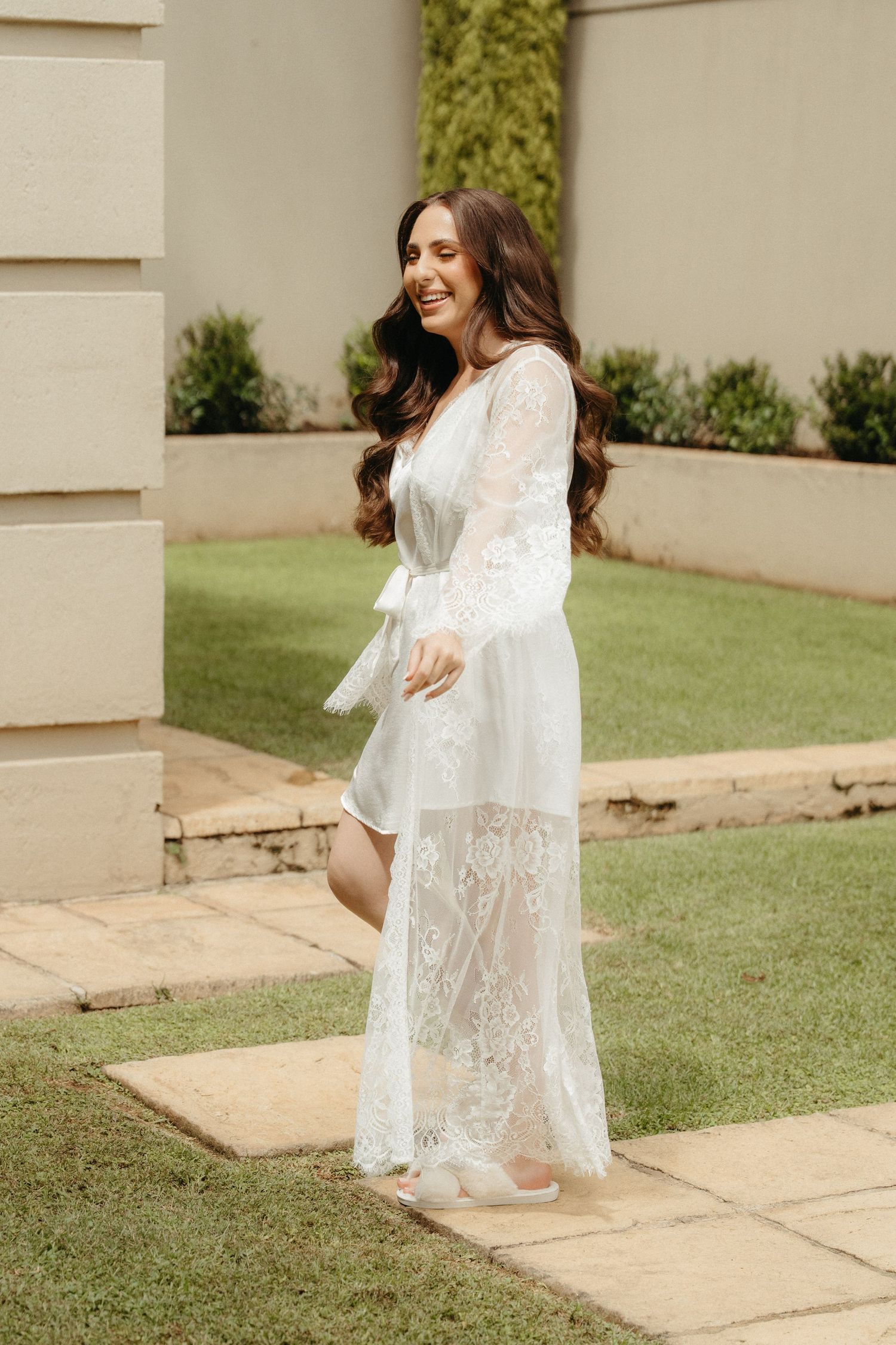 Woman in white lace bridal robe smiling outdoors on stone path with green garden background.