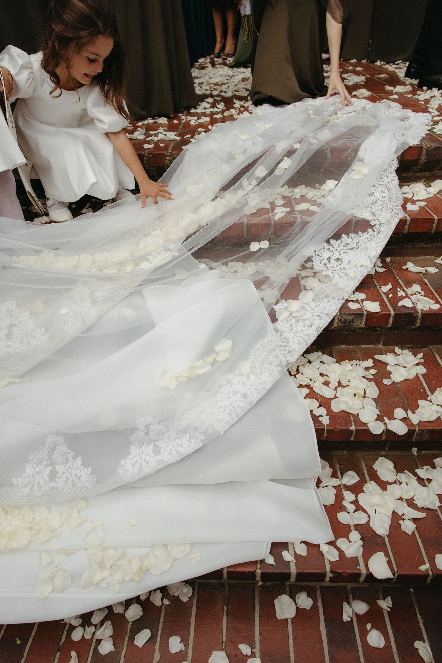 Flower girl arranging lace-trimmed bridal veil train on brick steps scattered with white rose petals.