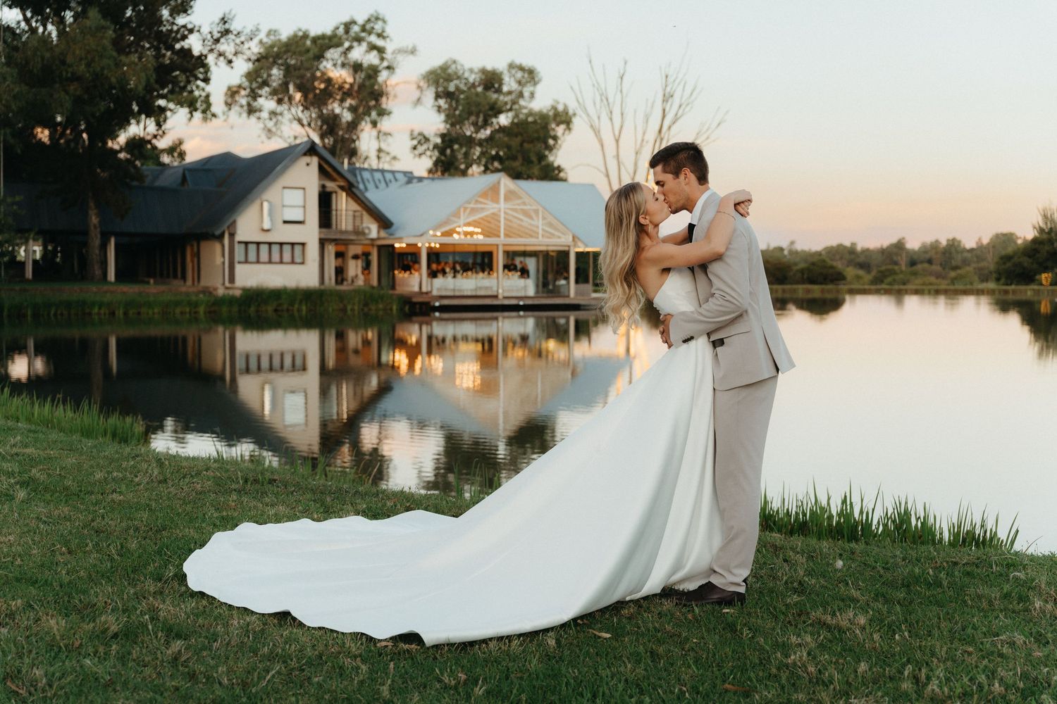 Bride and groom share a romantic kiss by a reflective pond at sunset, with a charming venue in the background.
