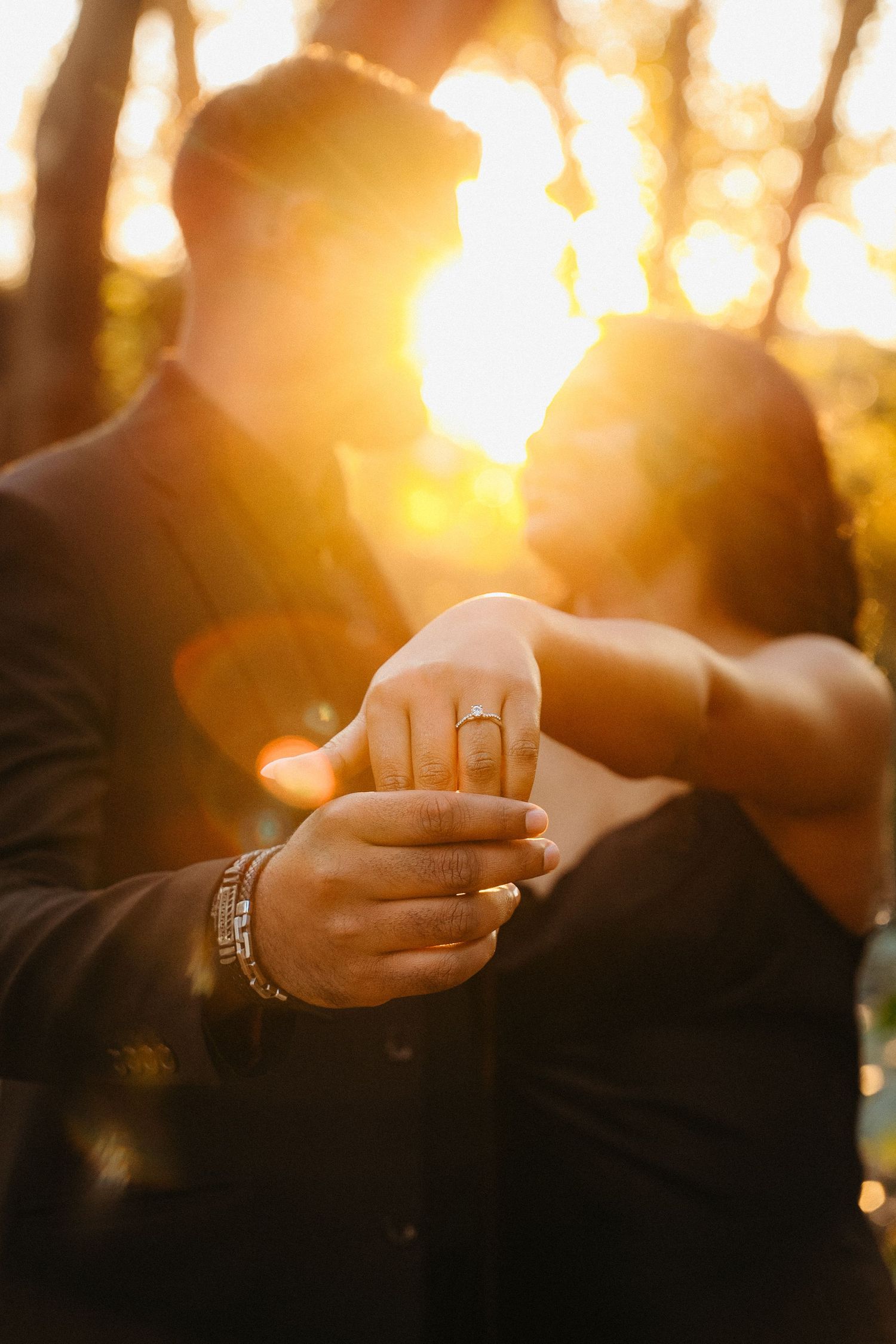 Couple showing off engagement ring at golden sunset, warm backlit glow highlighting diamond ring on her finger.