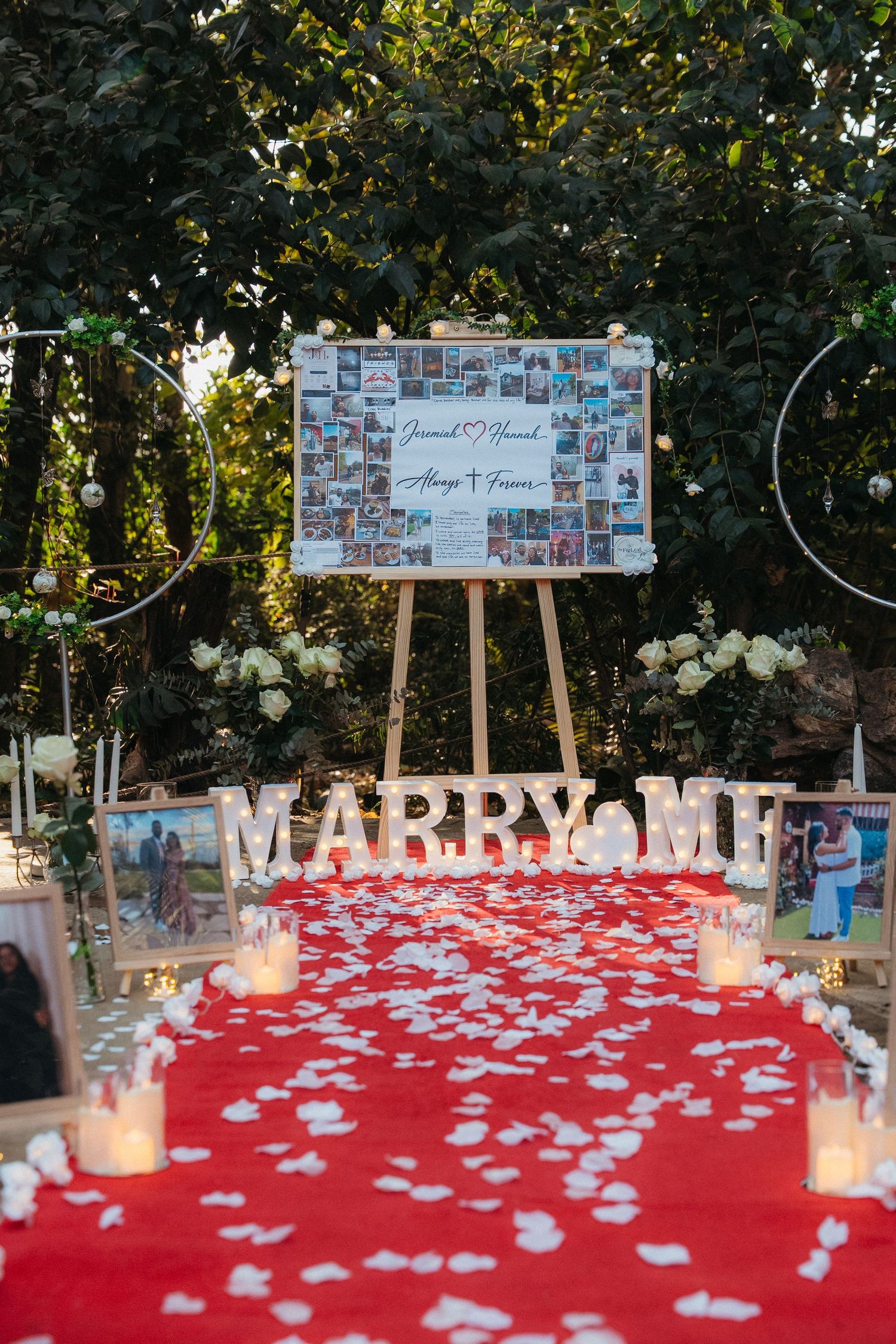 Romantic outdoor marriage proposal setup with red petal aisle, candles, MARRY ME letters, and photo collage board on easel.