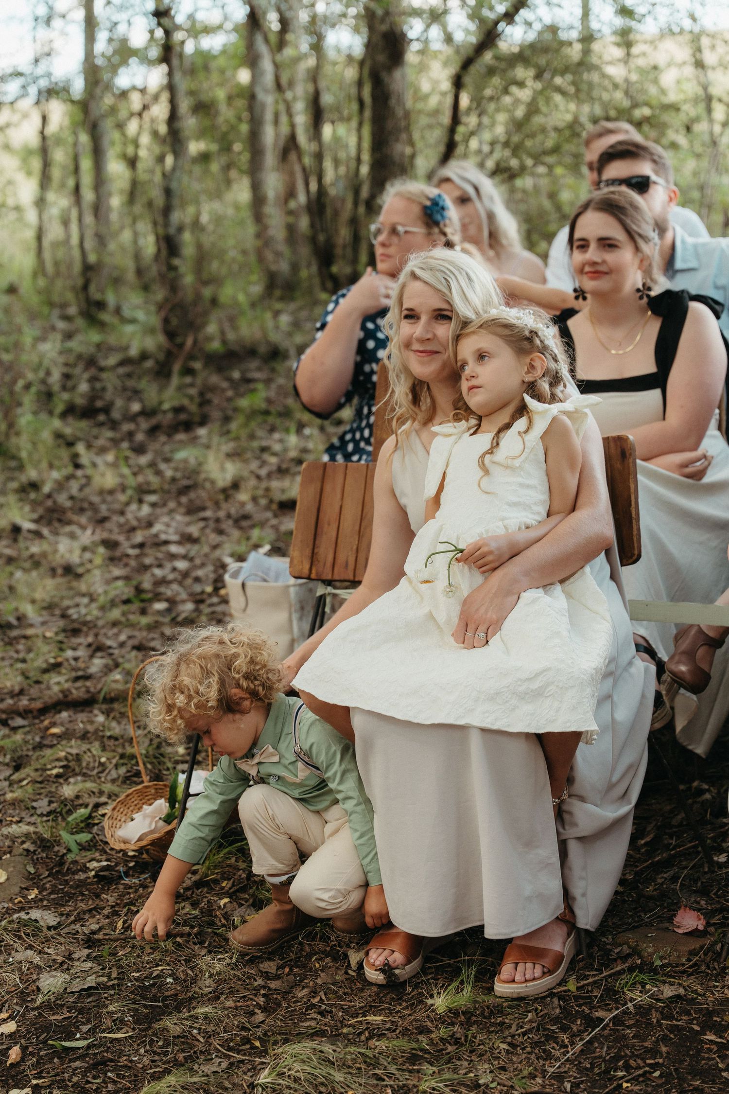 Bride in white dress sits with flower girl outdoors at woodland wedding ceremony with guests seated behind.