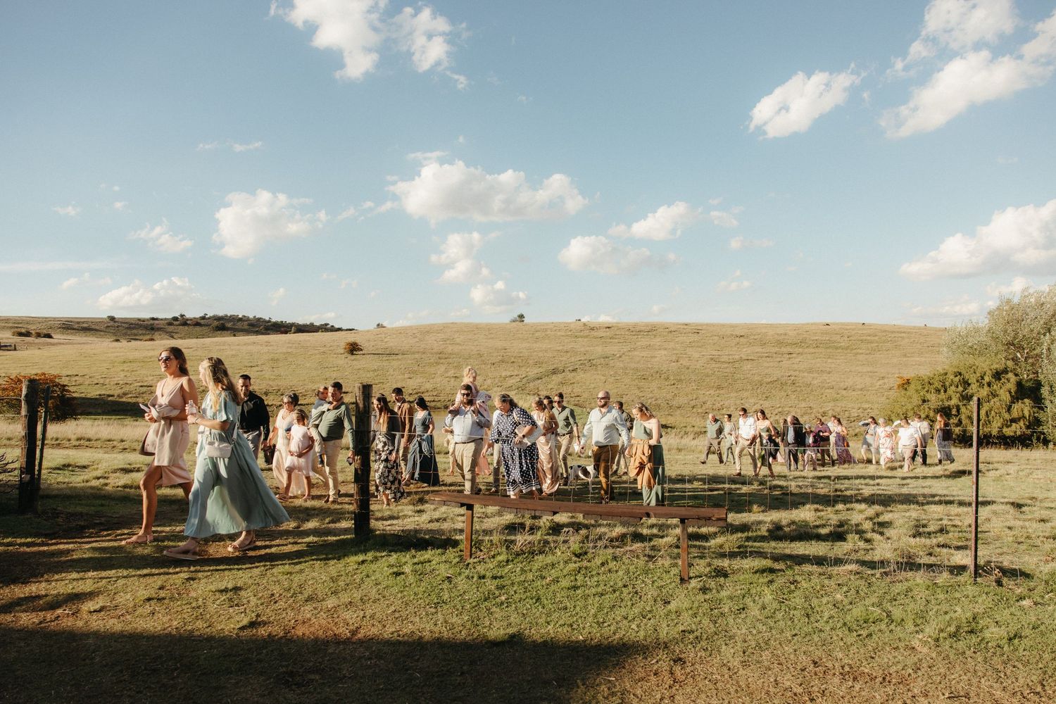 Guests gathered outdoors at a rustic farm wedding ceremony under a blue sky with rolling green fields.