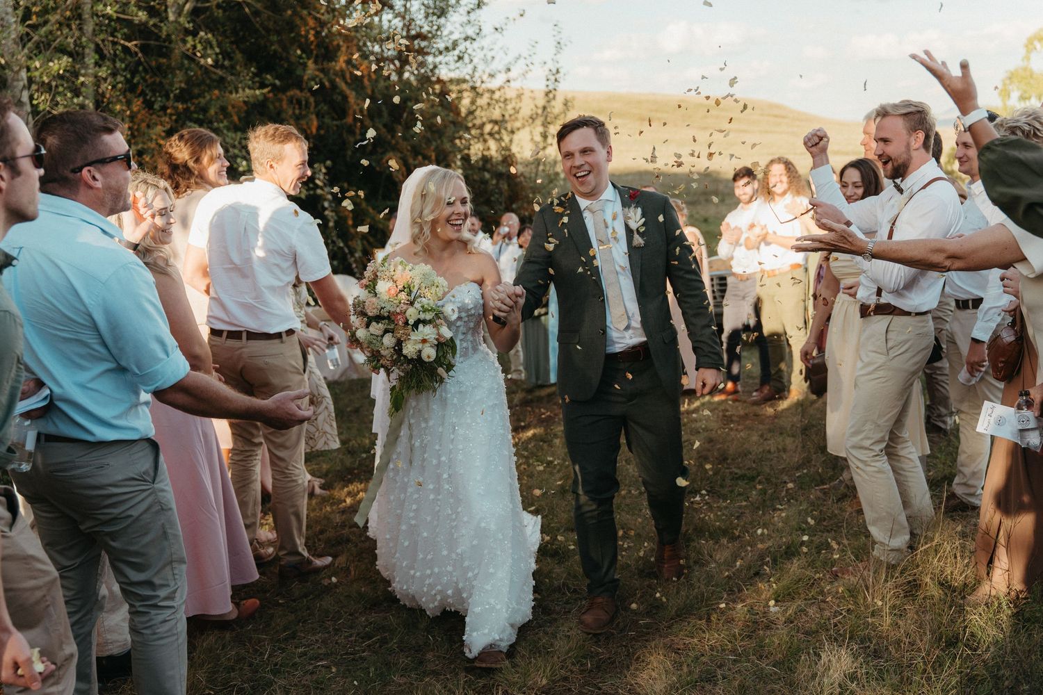 Bride and groom walk through cheering guests tossing confetti at outdoor wedding ceremony on sunny day.