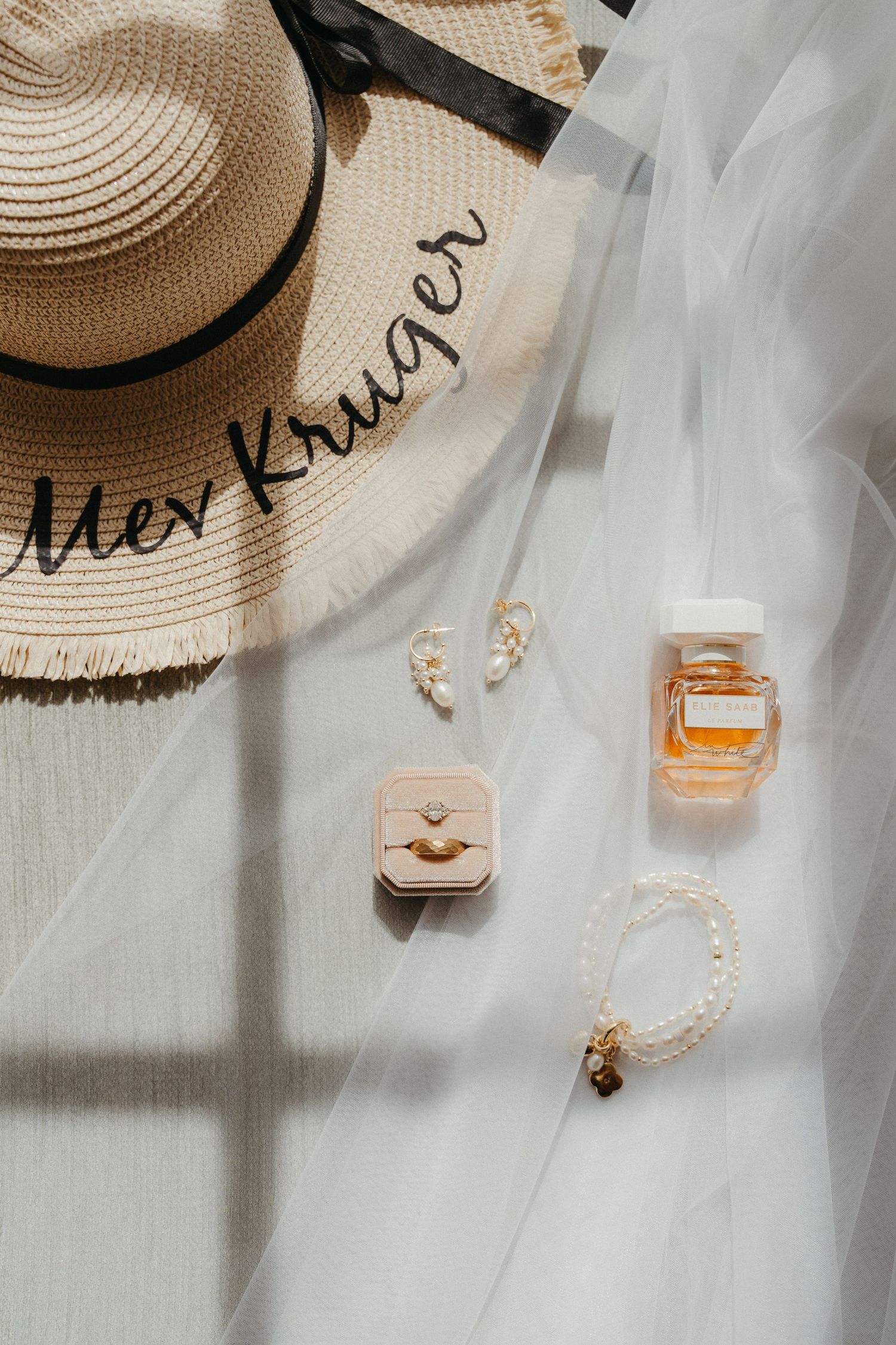 Bridal flat lay with personalized straw hat, gold ring box, pearl bracelet, and amber perfume bottle on white tulle.