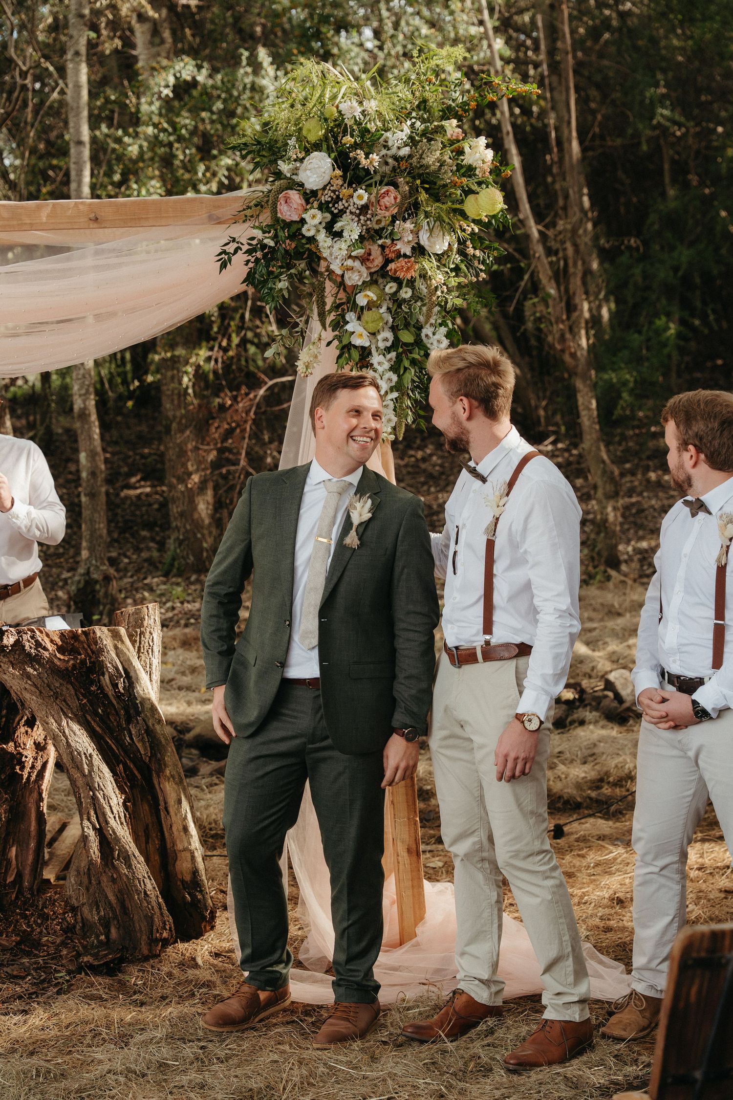 Groom in dark green suit smiling with groomsmen in suspenders at outdoor woodland wedding ceremony.