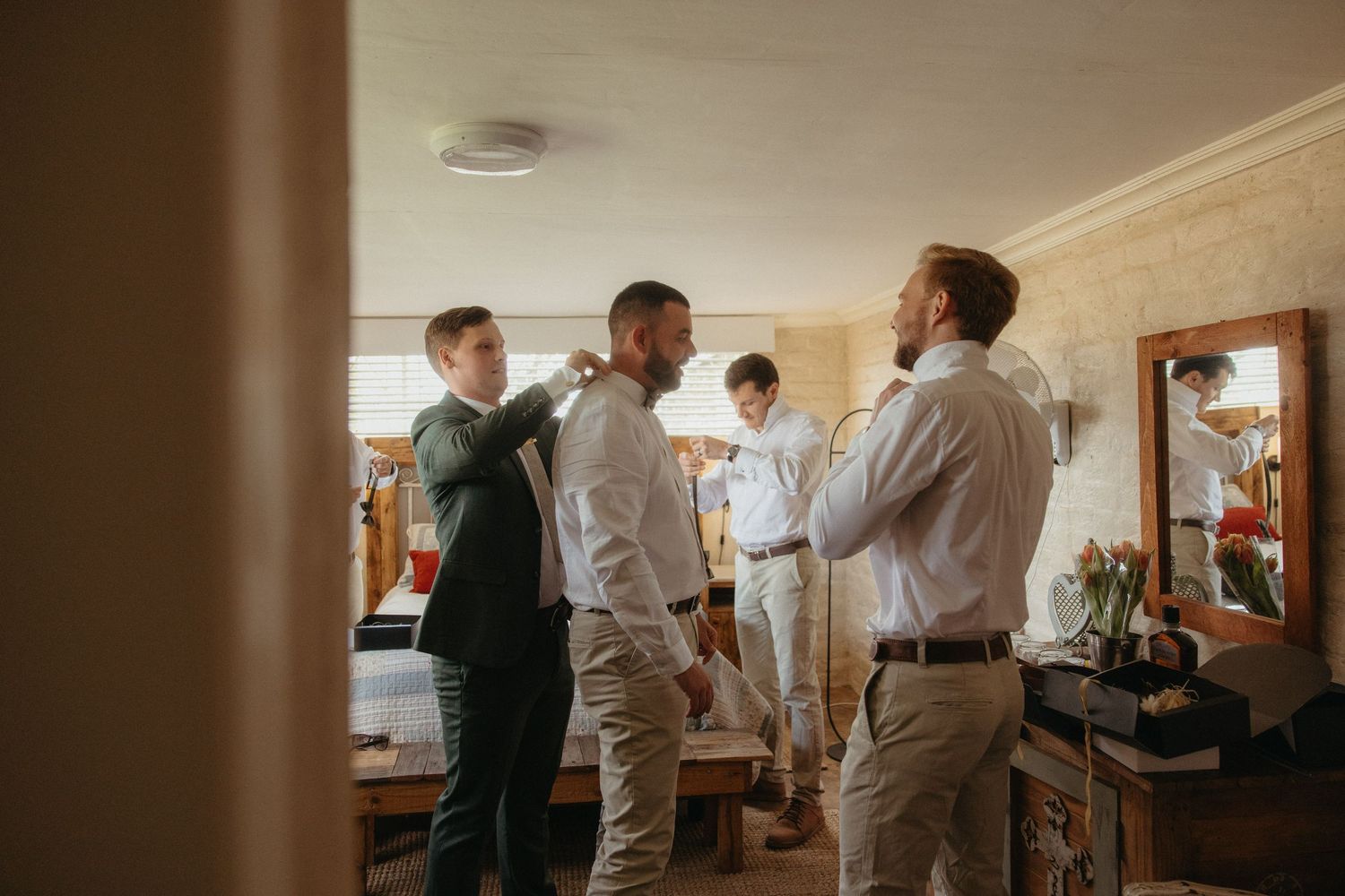 Groomsmen helping each other prepare for a wedding, adjusting suits in a warmly lit room.