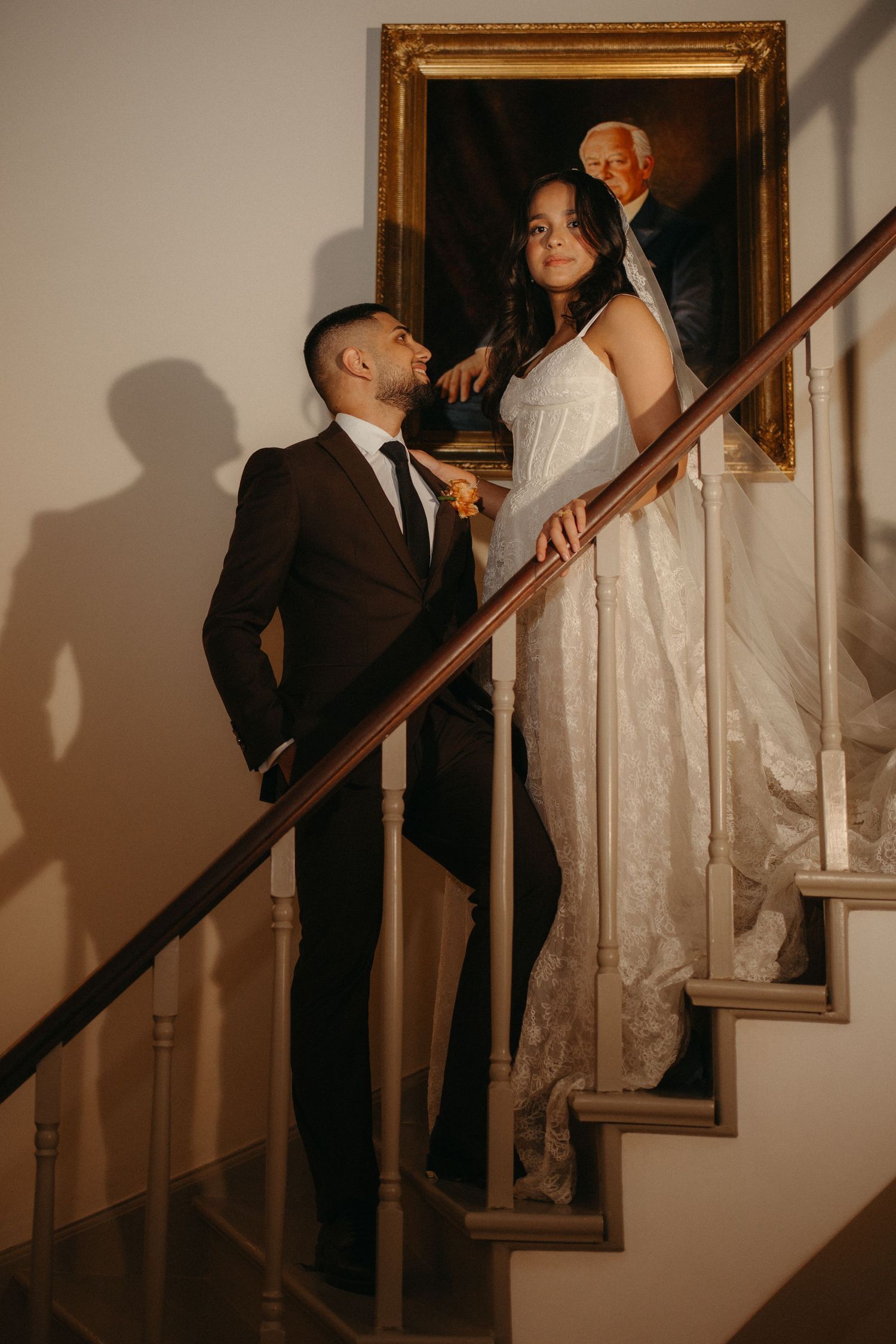 Bride and groom pose on elegant staircase beside gold-framed portrait, bride in white gown, groom in black suit.