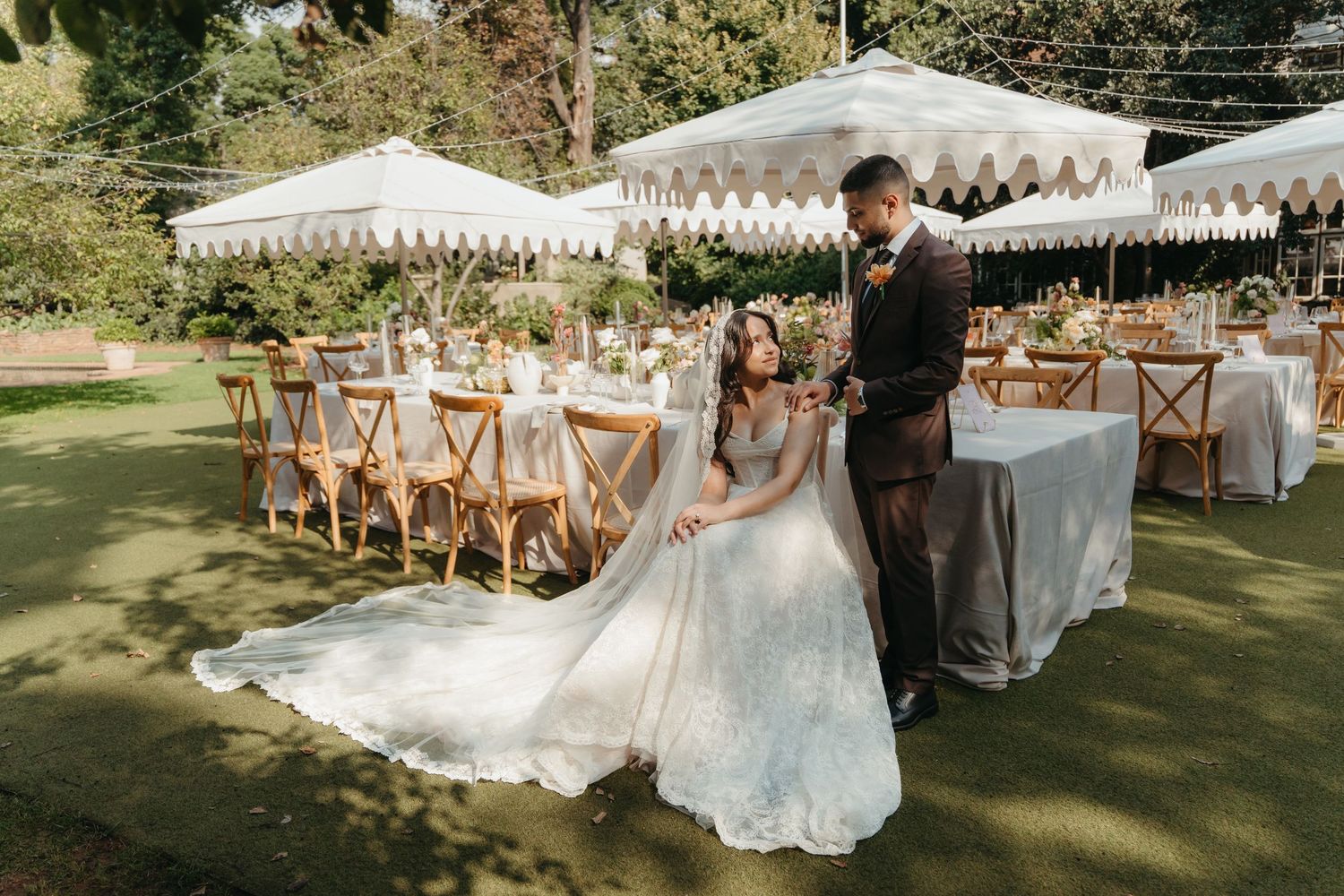 Bride and groom pose outdoors at elegant garden wedding reception with white marquee tents and wooden cross-back chairs.