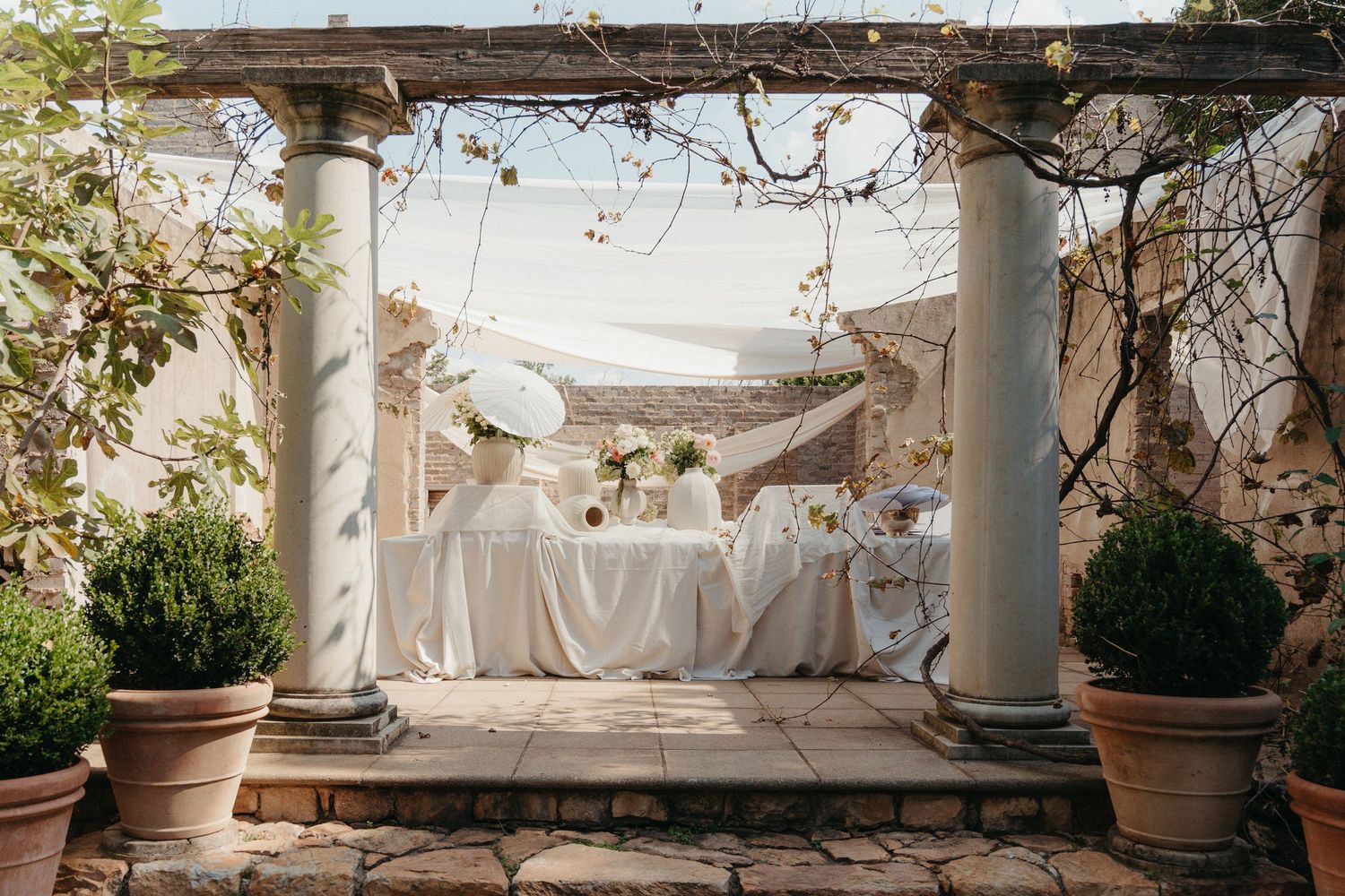 Elegant outdoor wedding table setup under a stone pergola with white linens, floral arrangements, and terracotta pots.
