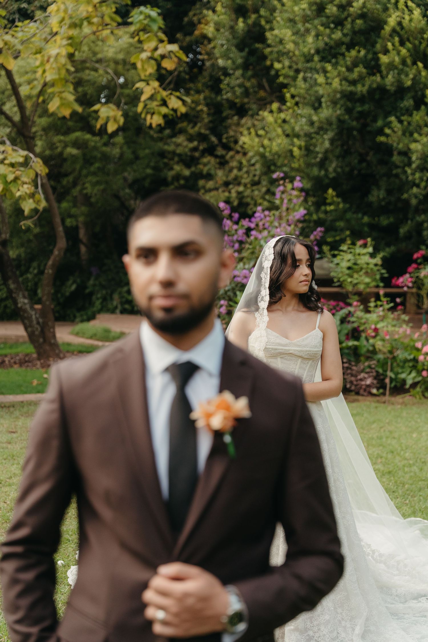 Bride in white gown and veil stands behind groom in brown suit with floral boutonniere in lush garden setting.