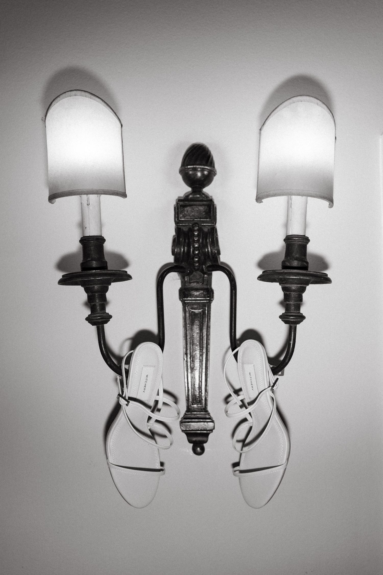 Black and white photo of white strappy sandals hanging on an ornate two-arm wall sconce with lamp shades.