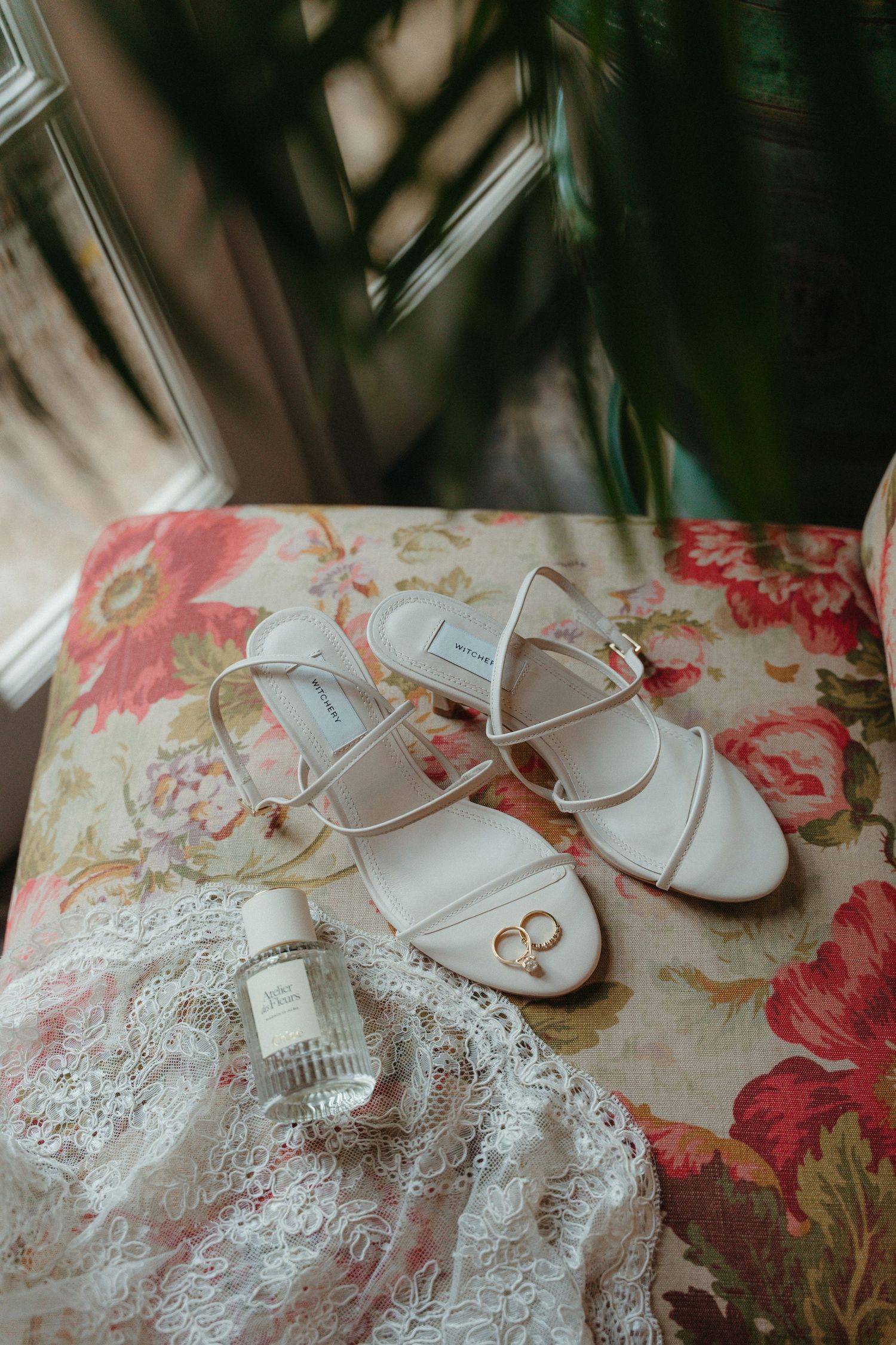 White strappy bridal heels with gold rings and perfume bottle on floral chair with lace fabric.