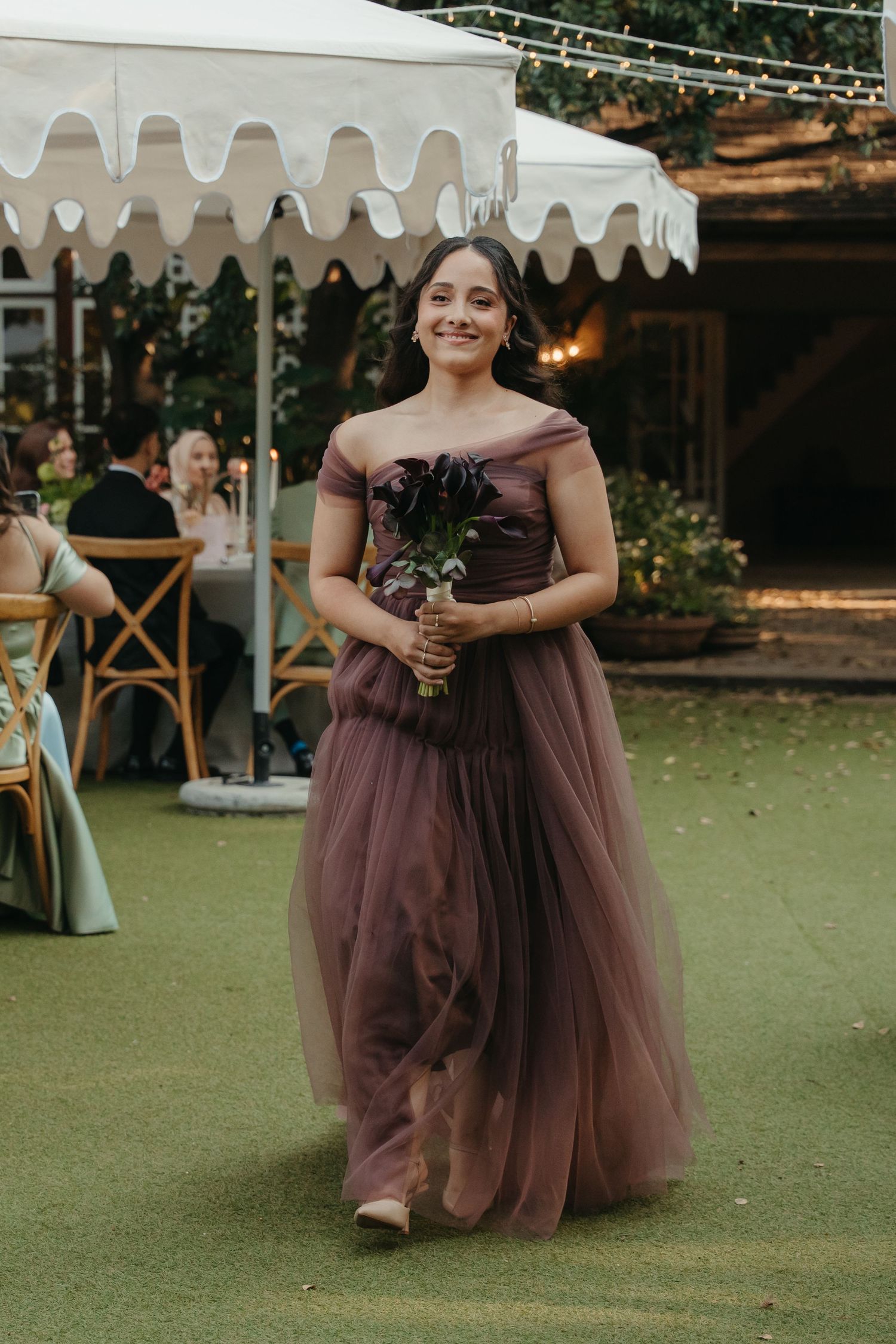 Bridesmaid in mauve off-shoulder tulle gown holding dark floral bouquet walks at outdoor wedding reception.