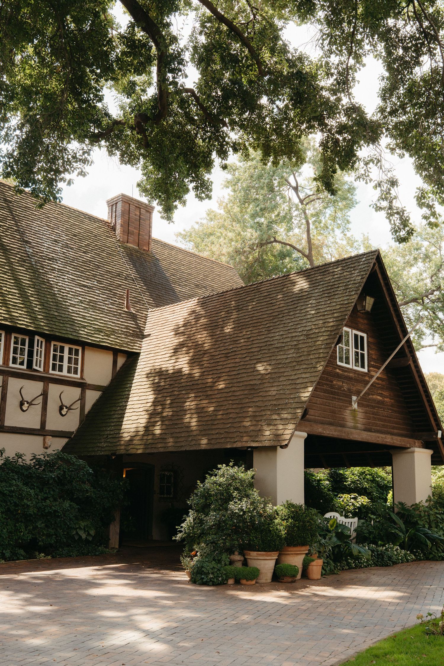 Beechwood Gardens Tudor-style estate with steep wood-shingled roof, brick chimney, and lush greenery framing a brick driveway entrance.