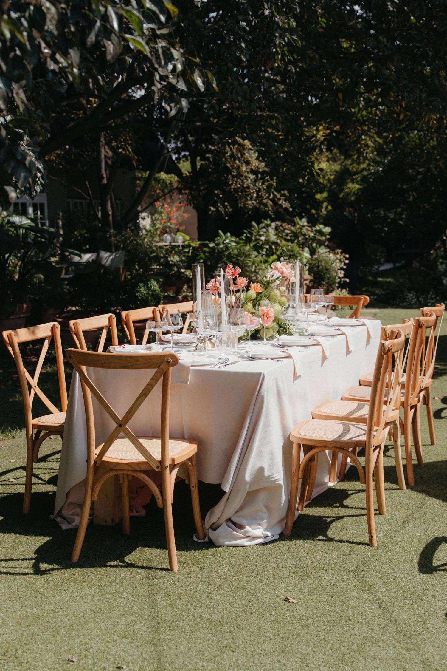 Elegant outdoor wedding reception table with white linen, floral centerpiece, and wooden cross-back chairs on green lawn.