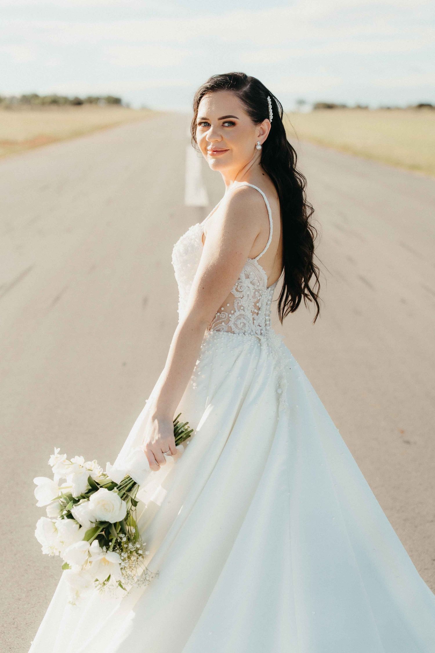 Bride in white lace ball gown holding bouquet of white flowers, standing on open road with long dark wavy hair.