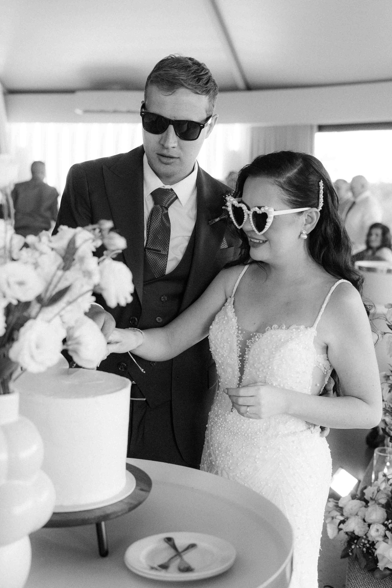 Bride and groom cutting wedding cake wearing sunglasses in black and white photo.